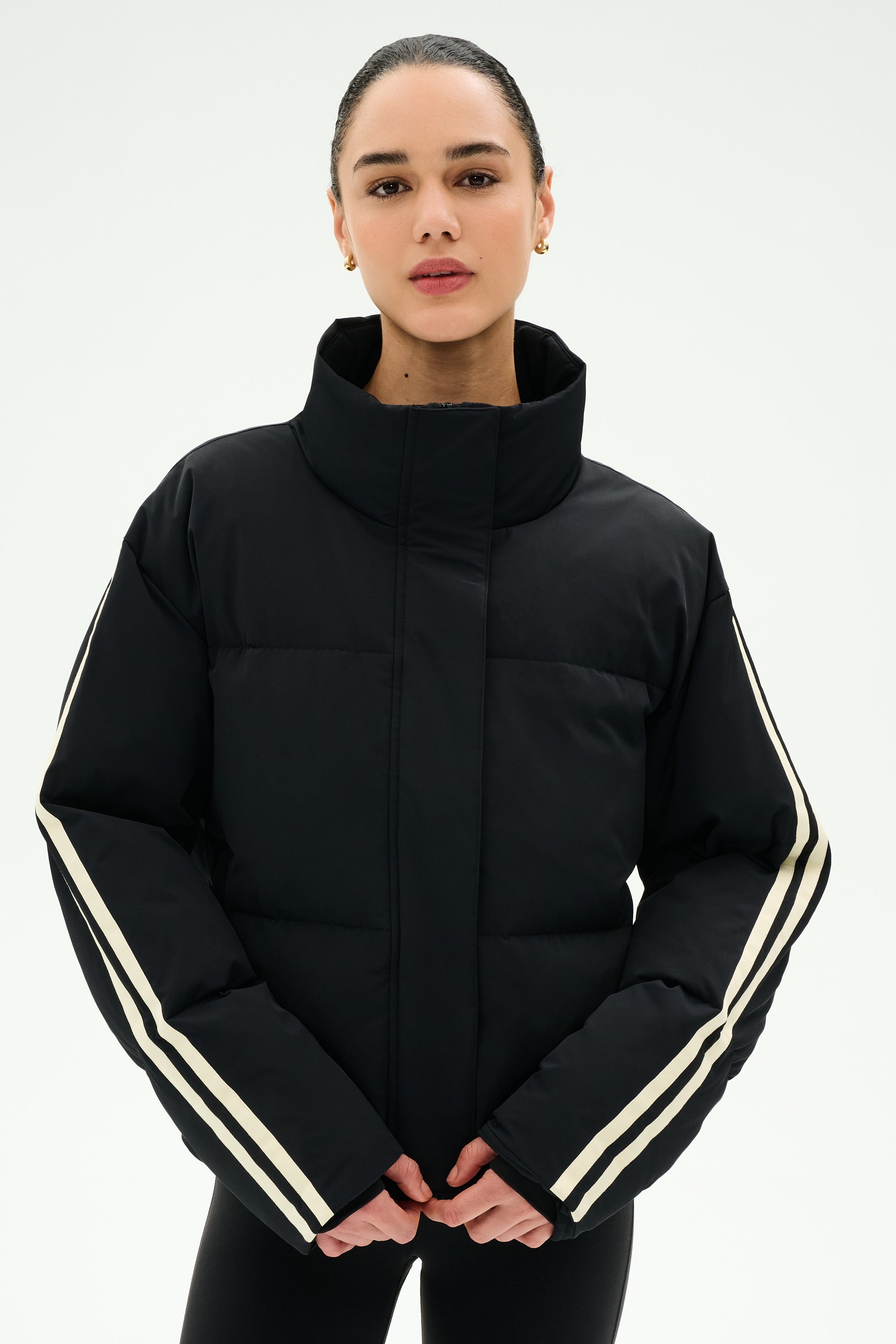 A person wearing the SPLITS59 Maeve Puffer Jacket in Black/Creme—crafted from 100% nylon and featuring white stripes on the sleeves—stands against a plain white background.