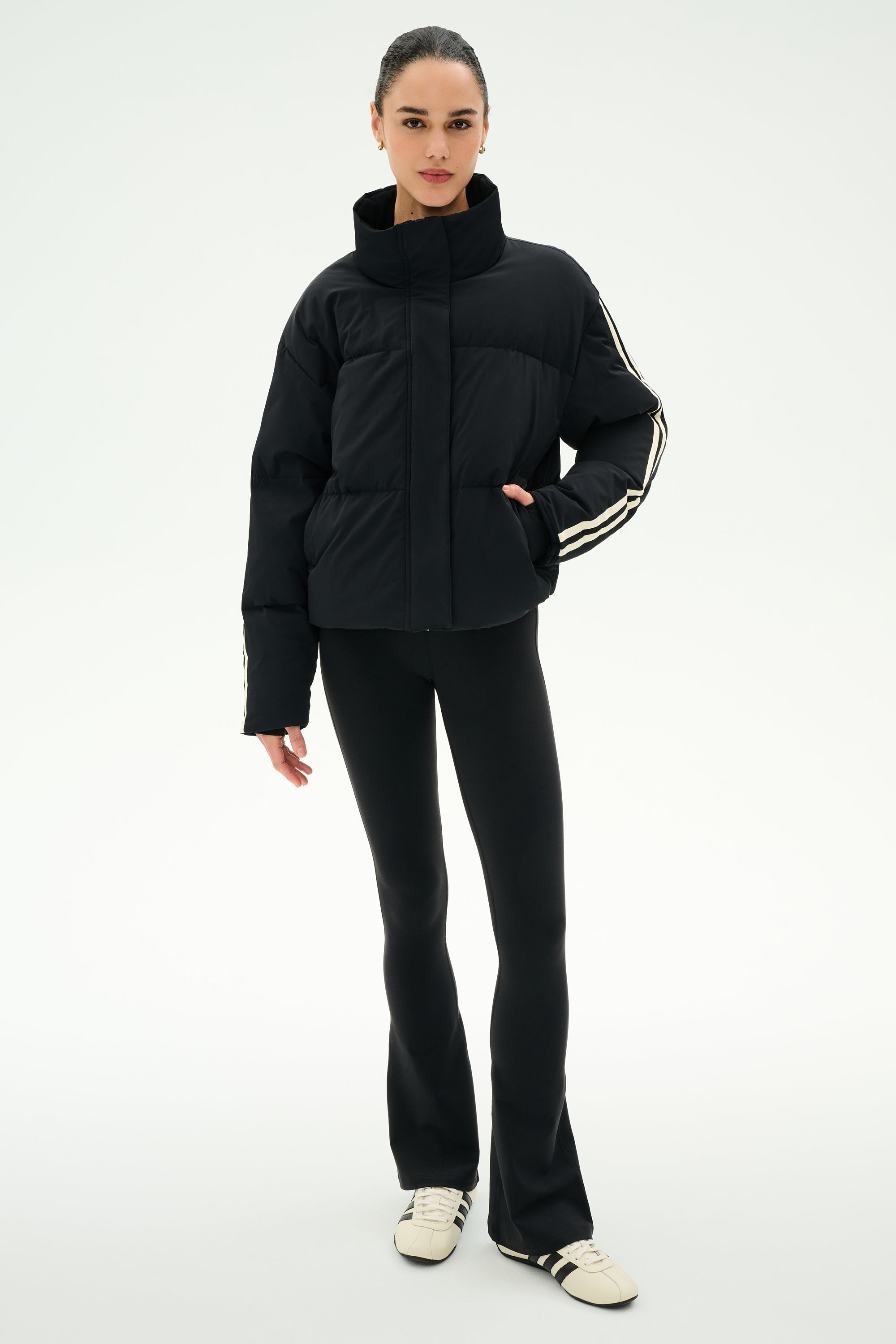 A person stands against a plain background wearing the SPLITS59 Maeve Puffer Jacket in Black/Creme, featuring 100% nylon construction and striped sleeves, paired with black pants and white sneakers with black stripes.