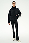 A person stands against a plain background wearing the SPLITS59 Maeve Puffer Jacket in Black/Creme, featuring 100% nylon construction and striped sleeves, paired with black pants and white sneakers with black stripes.