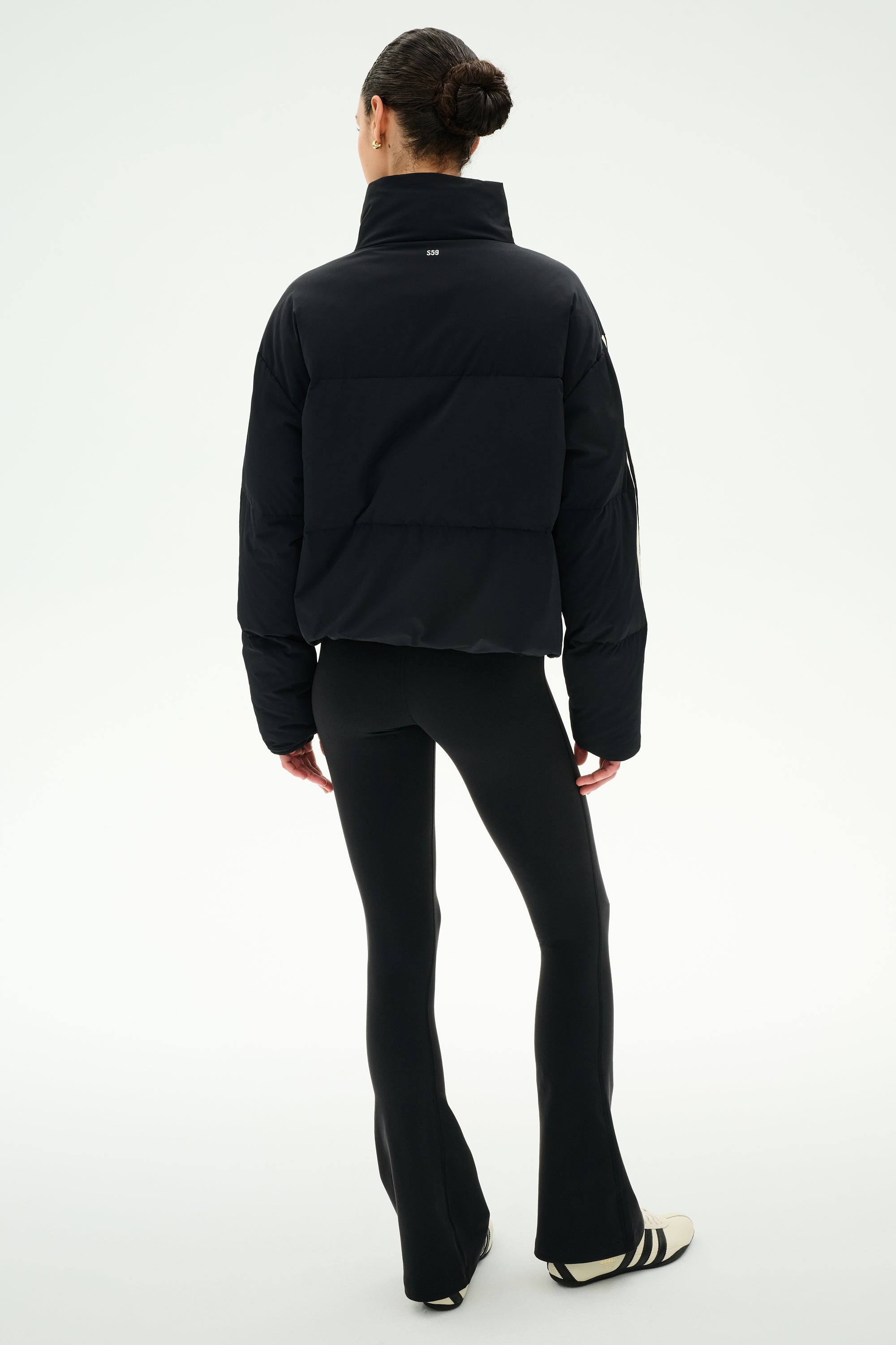 A person stands with their back to the camera, wearing the SPLITS59 Maeve Puffer Jacket in Black/Creme, paired with black flared leggings and black and white sneakers, set against a plain white background.