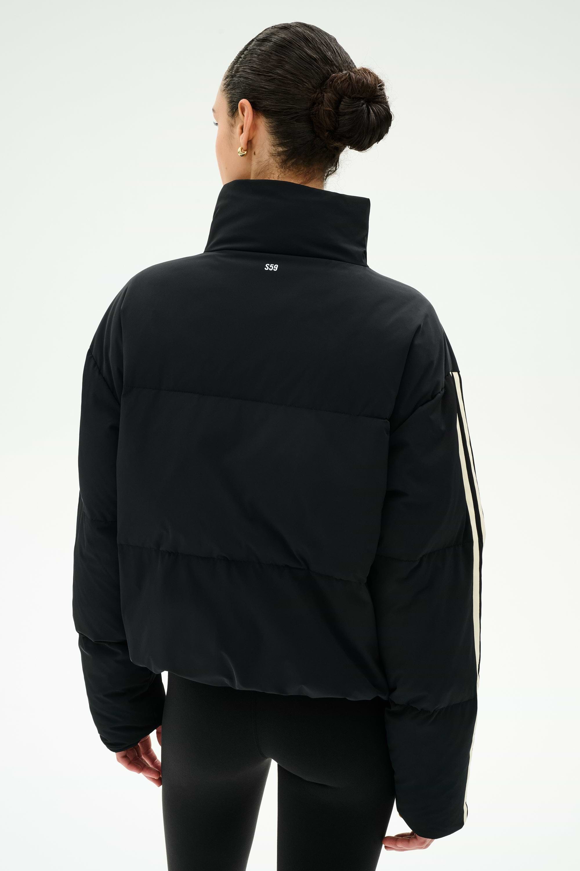 A woman with dark hair in a bun is photographed from behind against a plain white background, wearing the SPLITS59 Maeve Puffer Jacket in Black/Creme—crafted from 100% nylon—and black leggings.