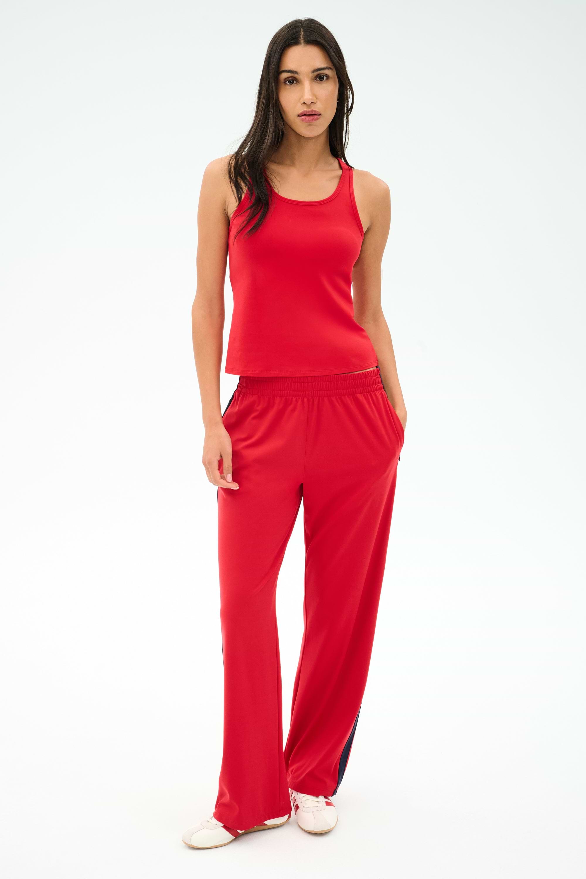 A woman faces forward in a SPLITS59 Mel Airweight Lite Tank in Pirate Red, paired with matching red pants with side stripes and white sneakers, set against a plain light backdrop.