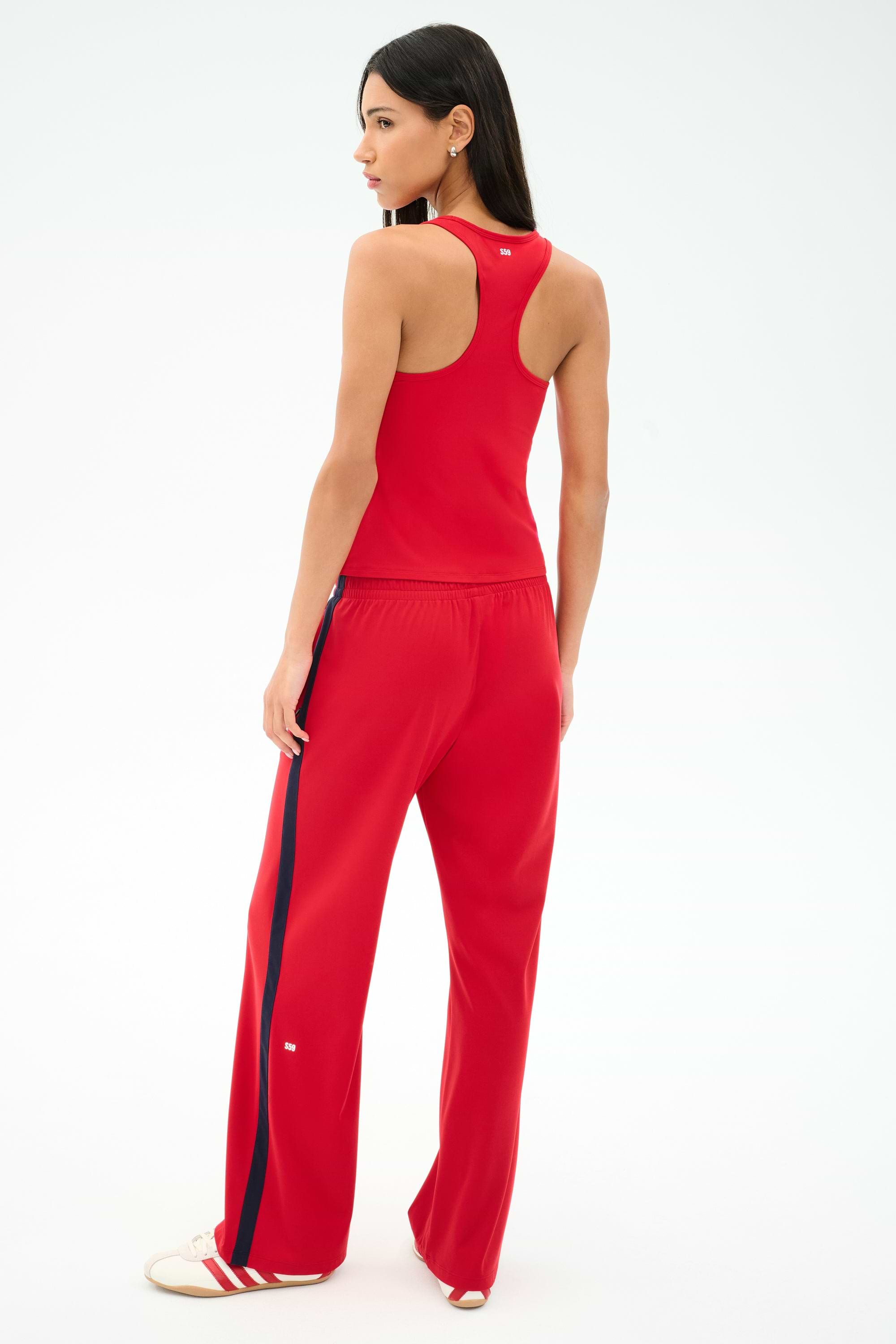 A woman faces away, wearing the SPLITS59 Mel Airweight Lite Tank in Pirate Red with matching track pants featuring a navy side stripe, paired with white sneakers.