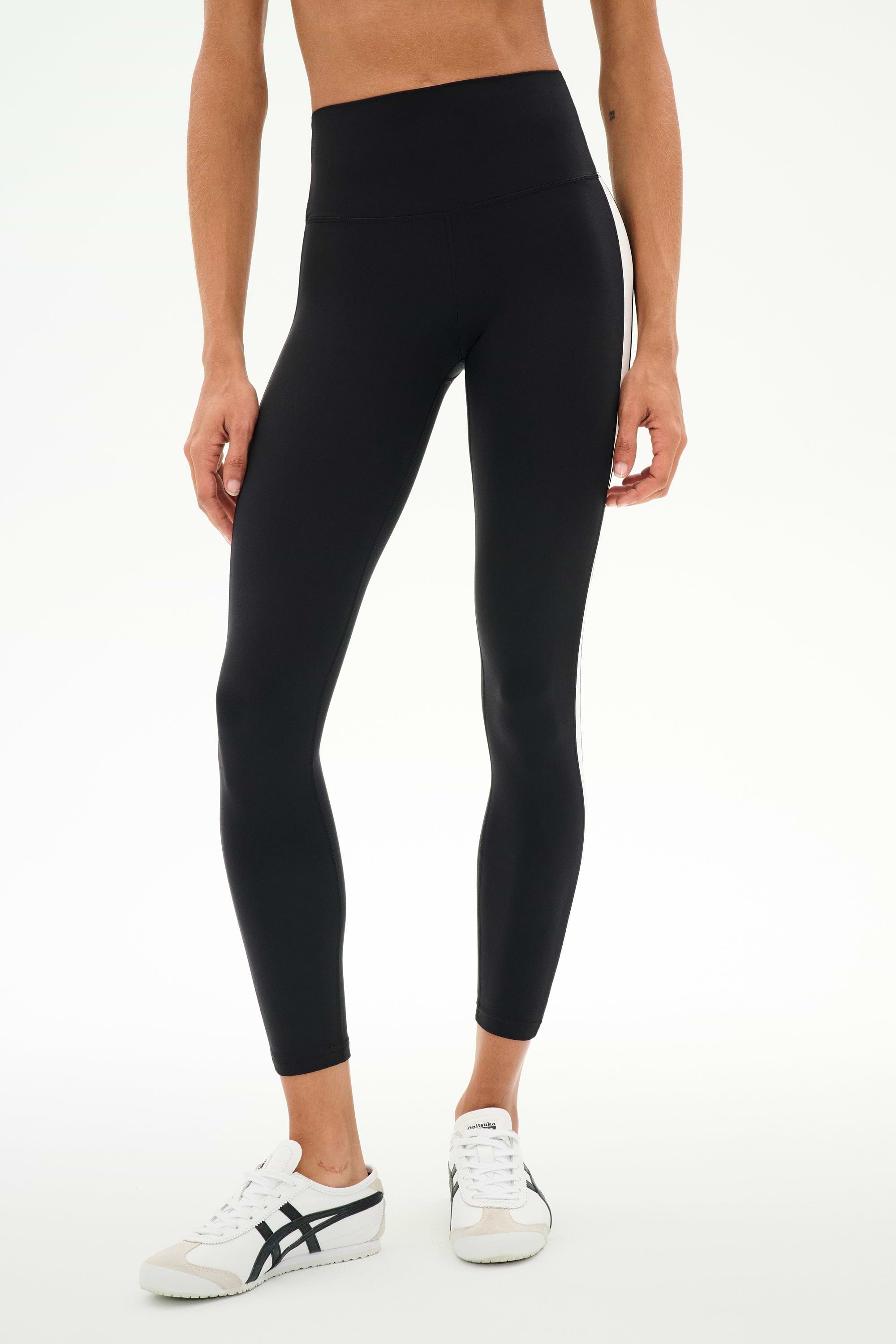 A person is seen wearing the SPLITS59 Clare High Waist Rigor 7/8 leggings in Black/White, paired with white sneakers and posed against a plain white background. Only the lower torso and legs are visible, highlighting these rigor-fabric high-waist leggings as an ideal option for gym workouts.