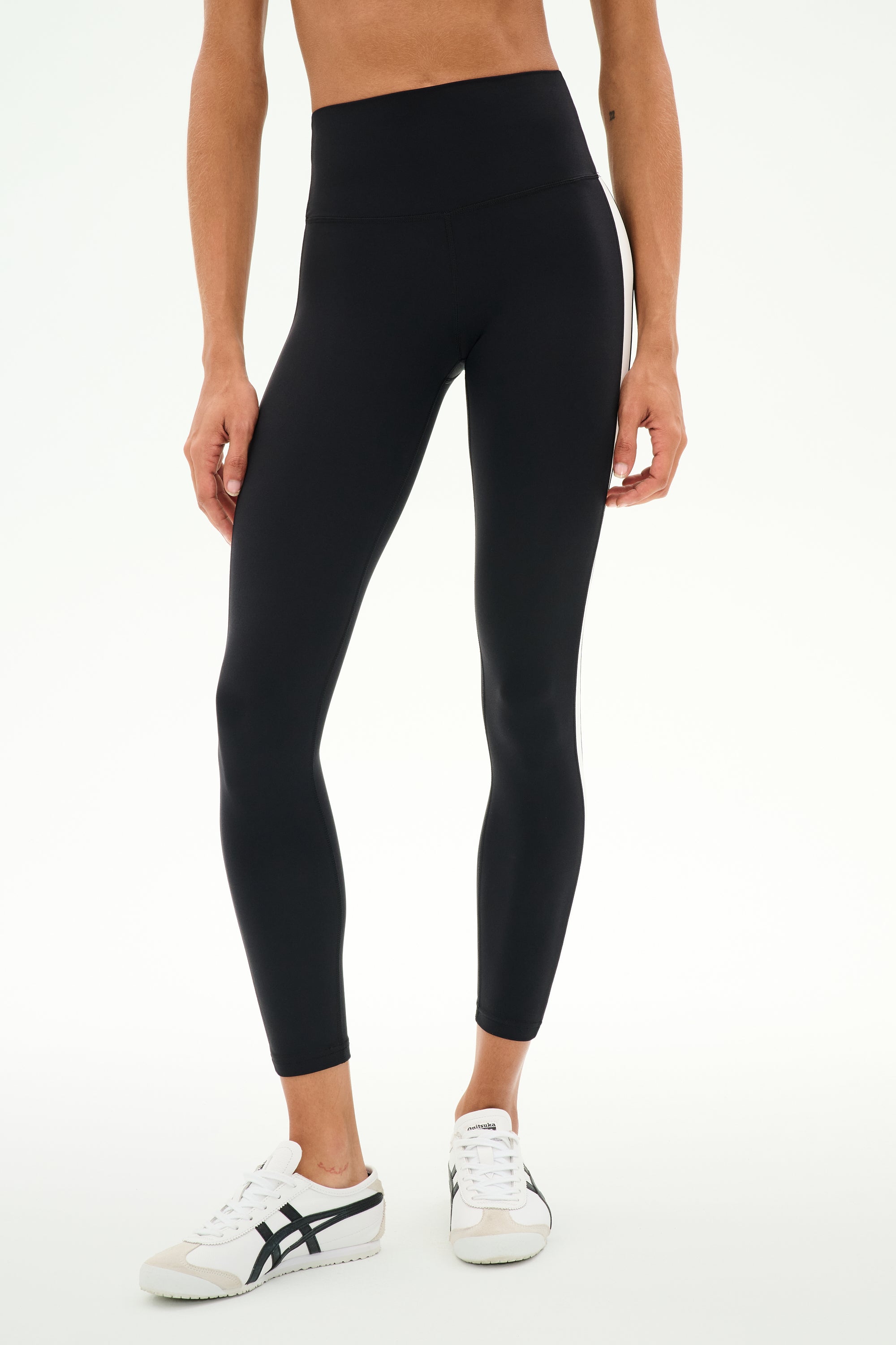 A person is seen wearing the SPLITS59 Clare High Waist Rigor 7/8 leggings in Black/White, paired with white sneakers and posed against a plain white background. Only the lower torso and legs are visible, highlighting these rigor-fabric high-waist leggings as an ideal option for gym workouts.