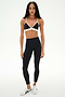 A woman stands facing forward against a plain white background, wearing the SPLITS59 Sage Rigor Bra in Black/White, paired with black leggings and white sneakers.