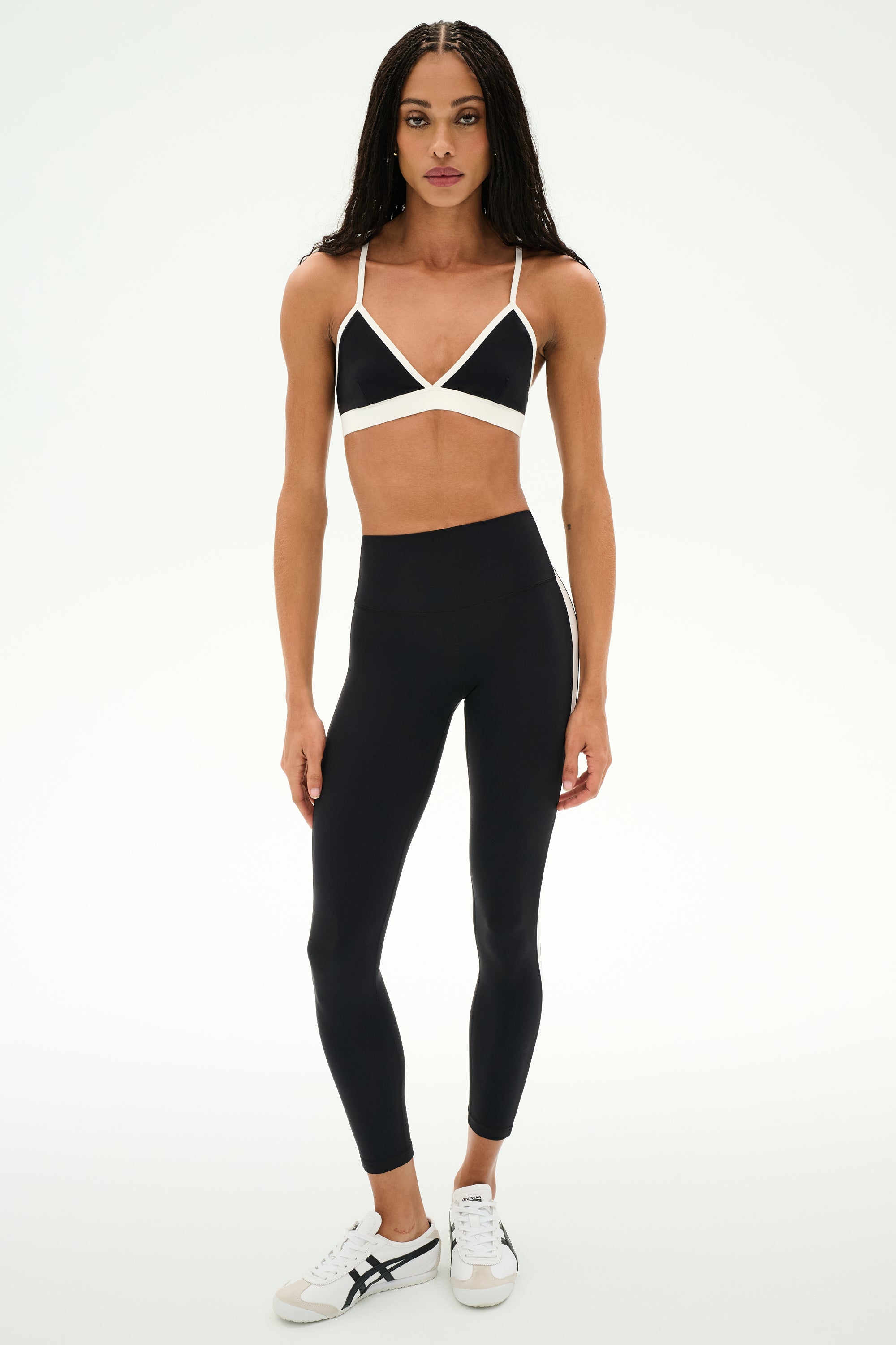 A woman stands facing forward against a plain white background, wearing the SPLITS59 Sage Rigor Bra in Black/White, paired with black leggings and white sneakers.