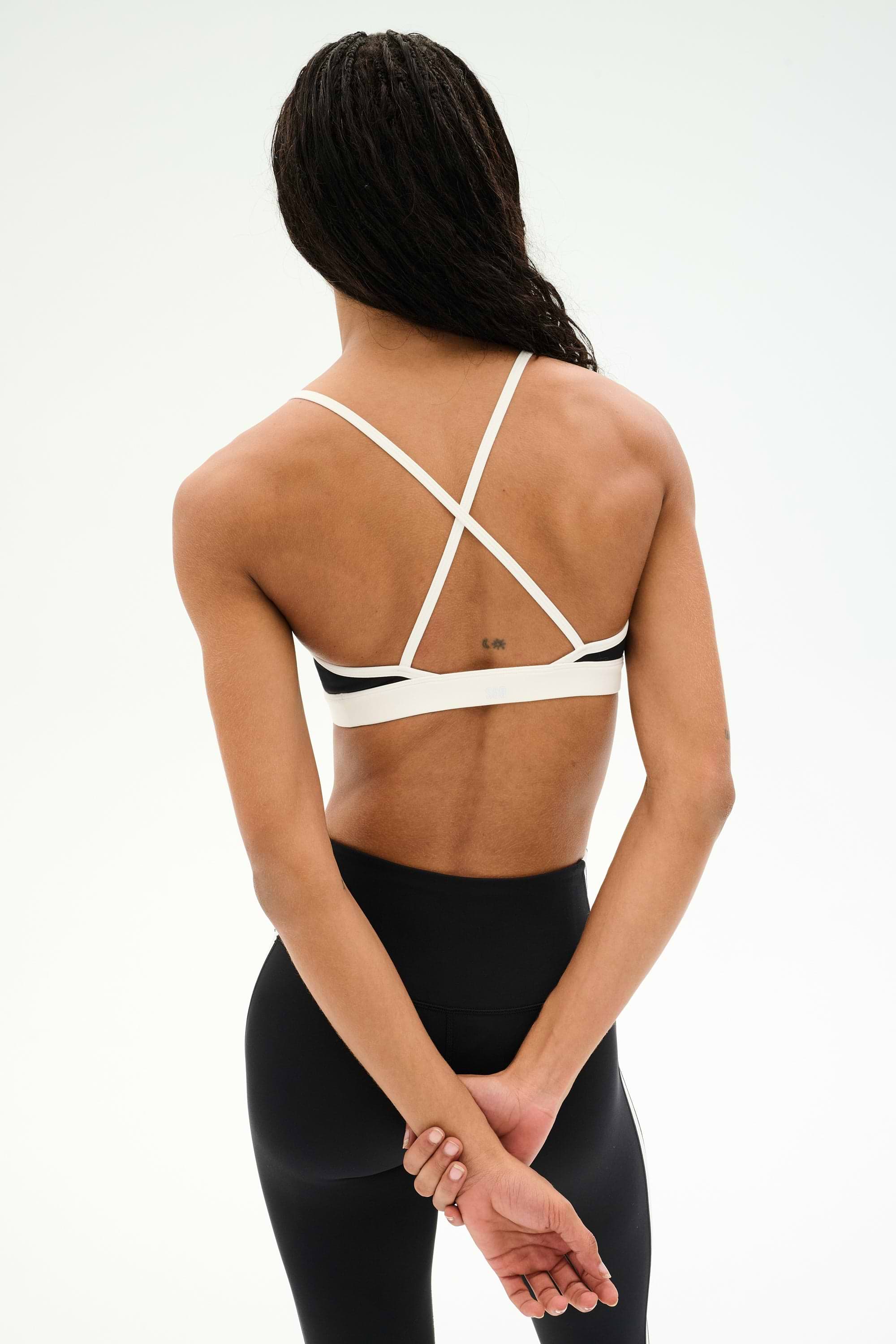 A person stands with their back to the camera, wearing the SPLITS59 Sage Rigor Bra in Black/White and black nylon-spandex leggings. Their hands are clasped behind their back.