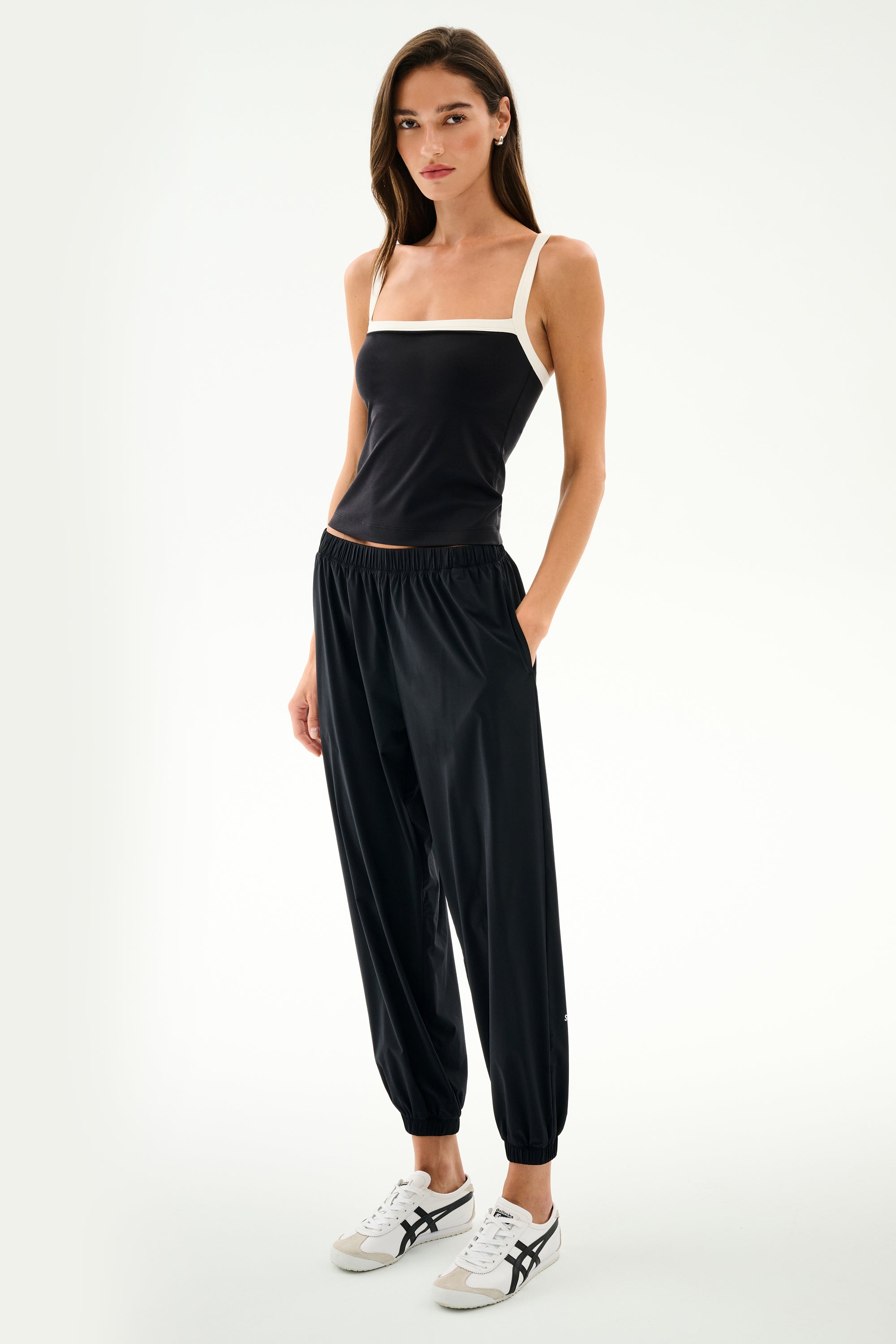 A woman wears the SPLITS59 Monah Rigor Tank in Black/White, featuring contrasting white straps and crafted from Rigor fabric, styled with black jogger pants and white sneakers against a plain white background—ideal for gym sessions or casual outings.