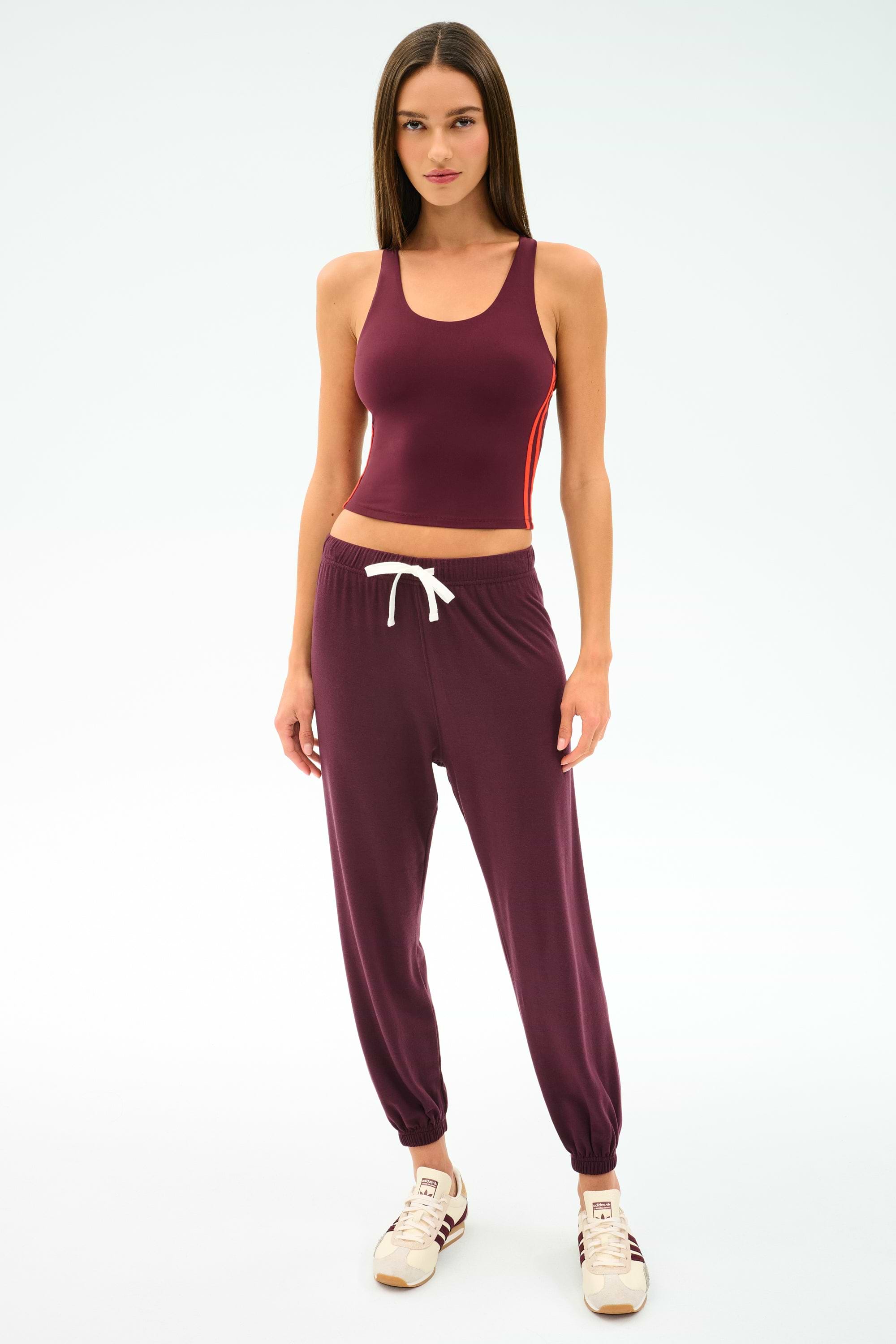 A woman faces forward in the SPLITS59 Ella Airweight Tank in Dark Cherry/Valencia, paired with matching joggers featuring a white drawstring and beige sneakers, all set against a plain white background.
