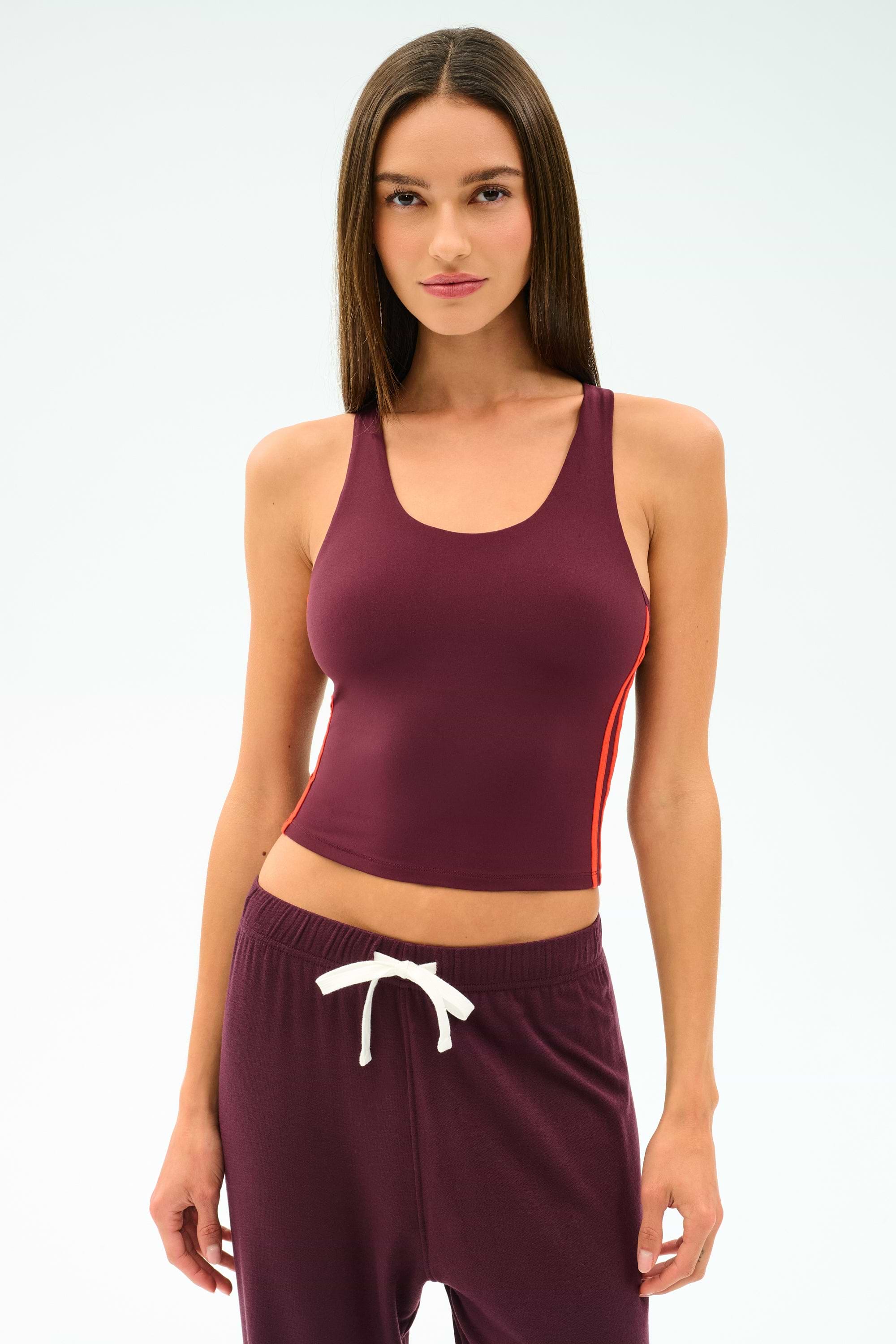 A woman stands against a plain light background wearing the SPLITS59 Ella Airweight Tank in Dark Cherry/Valencia, paired with matching sweatpants featuring a white drawstring, both crafted from stretchy nylon spandex fabric.