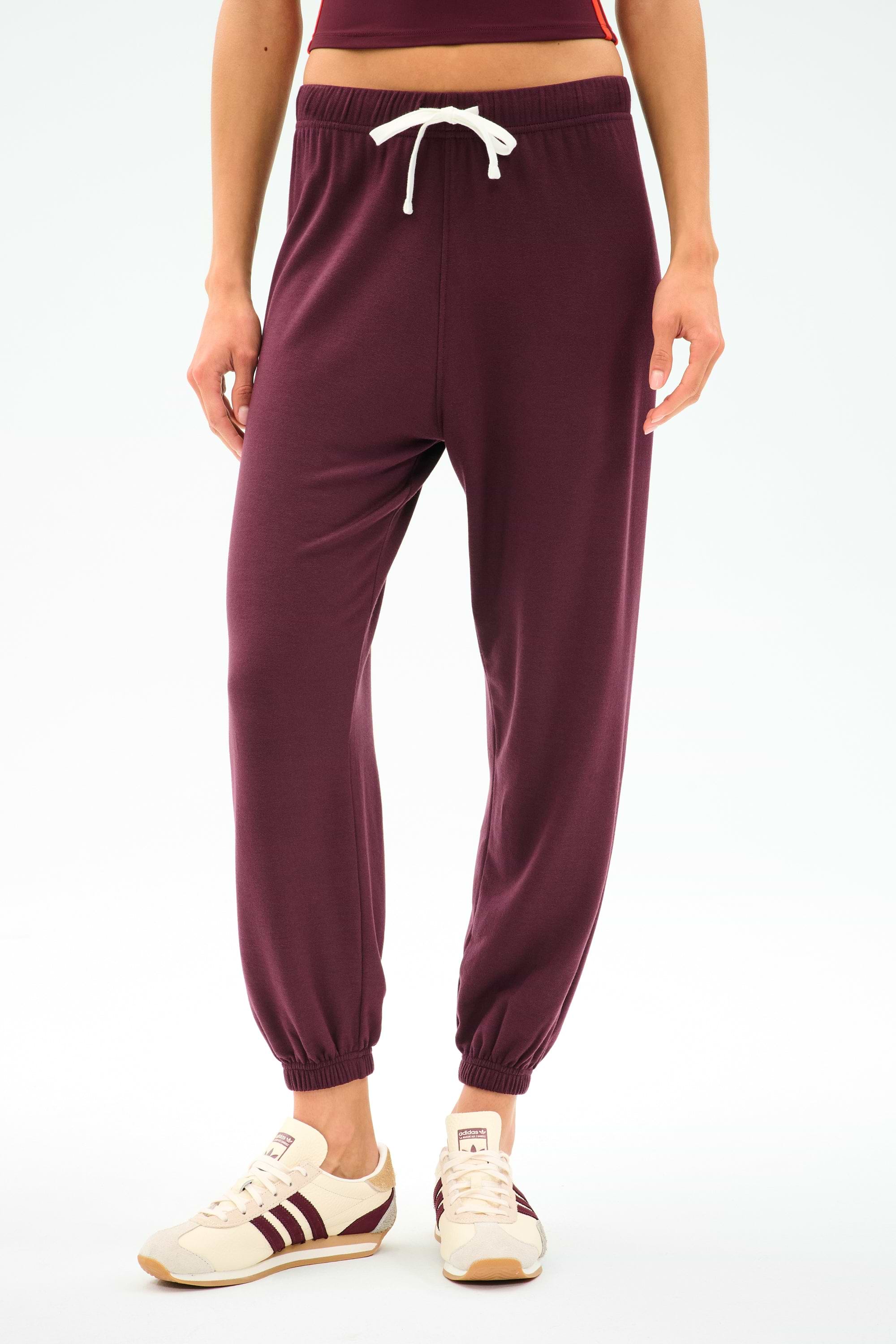A person is wearing SPLITS59's Andie Oversized Fleece Sweatpant in Dk Cherry, styled with beige sneakers featuring maroon stripes, standing against a plain white background.