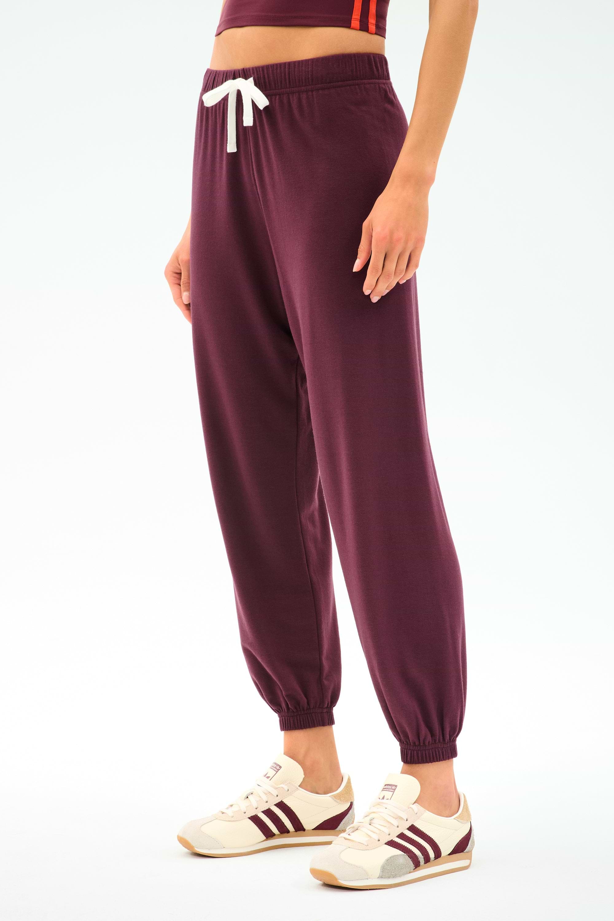 A person wears SPLITS59’s Andie Oversized Fleece Sweatpant in Dk Cherry with a white drawstring, paired with a cropped top and white sneakers featuring burgundy stripes, standing against a plain background.