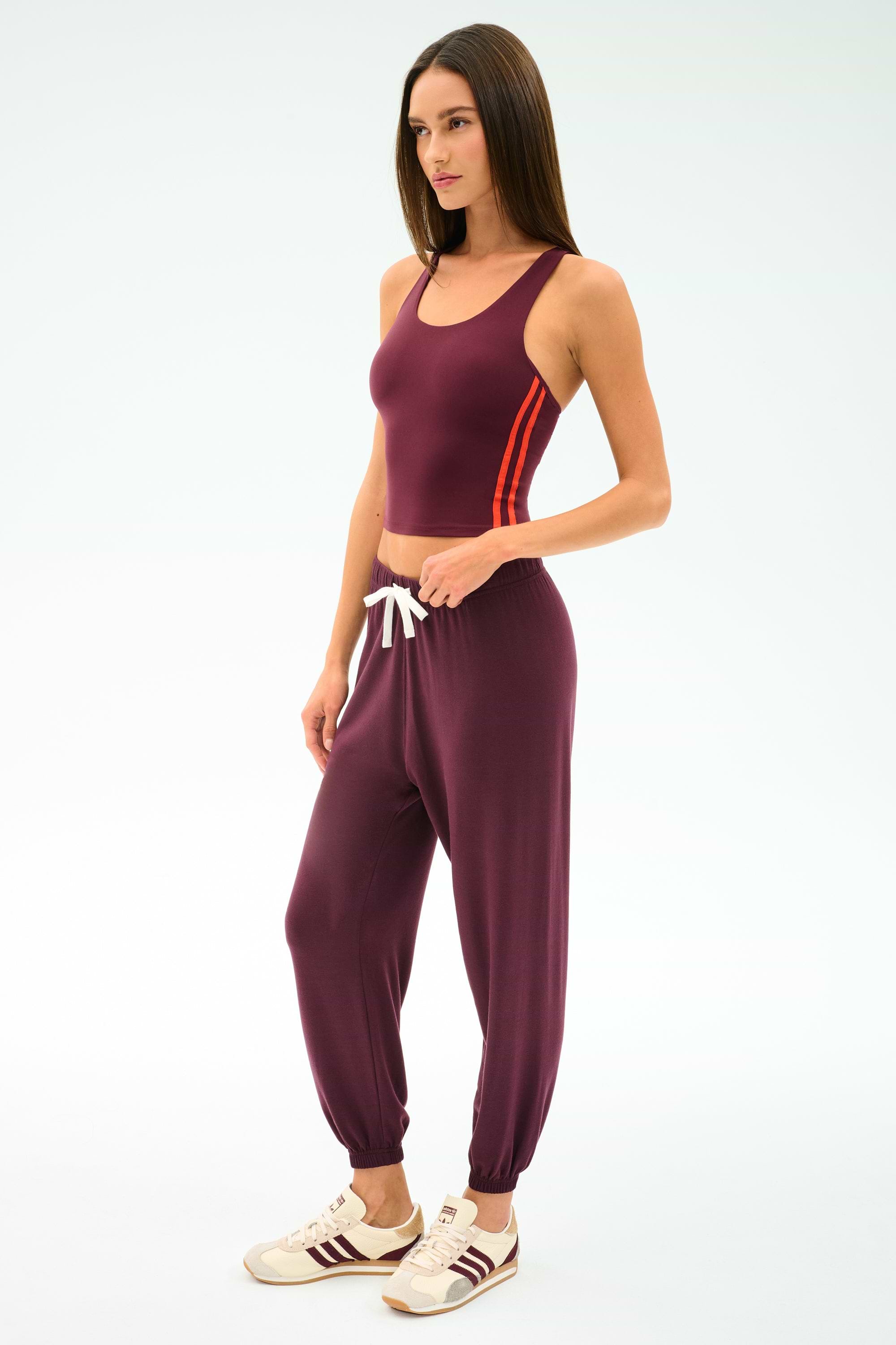 A woman wears the SPLITS59 Ella Airweight Tank in Dark Cherry/Valencia with matching jogger pants and white sneakers, standing against a plain light background.
