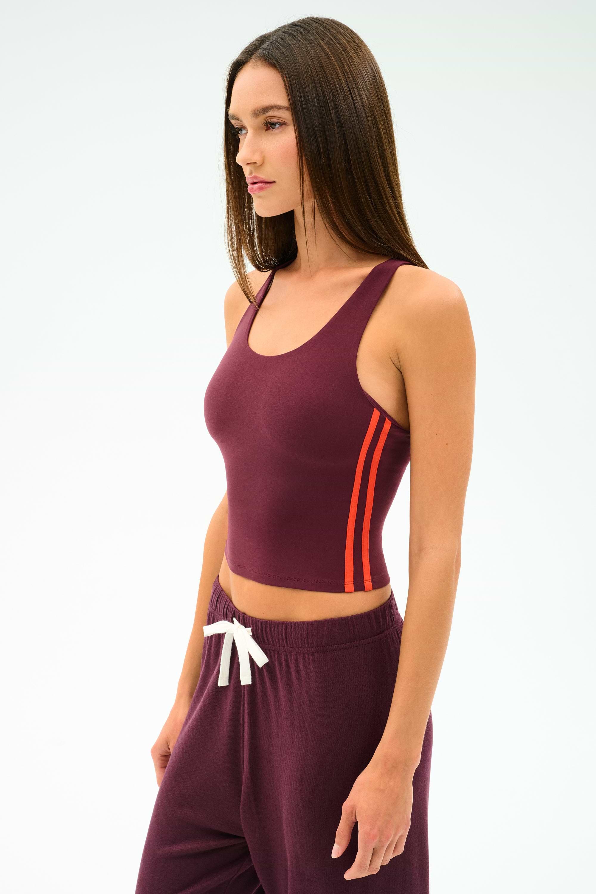A woman wears the SPLITS59 Ella Airweight Tank in Dark Cherry/Valencia, featuring orange side stripes and matching pants with a white drawstring, made from stretchy nylon spandex, against a plain light background.