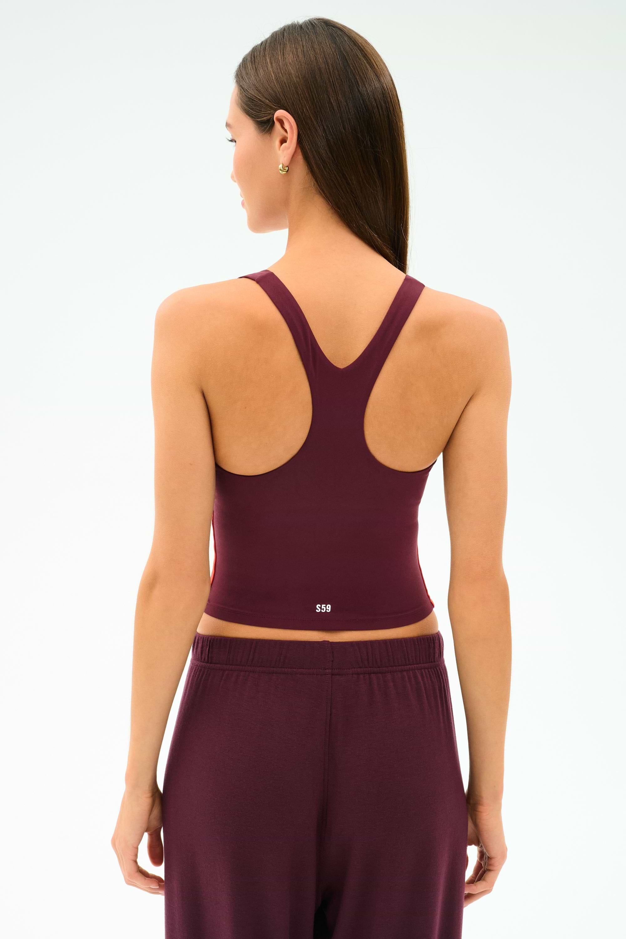 A woman with straight brown hair is seen from the back, wearing the SPLITS59 Ella Airweight Tank in Dark Cherry/Valencia, paired with matching pants, against a plain light background.