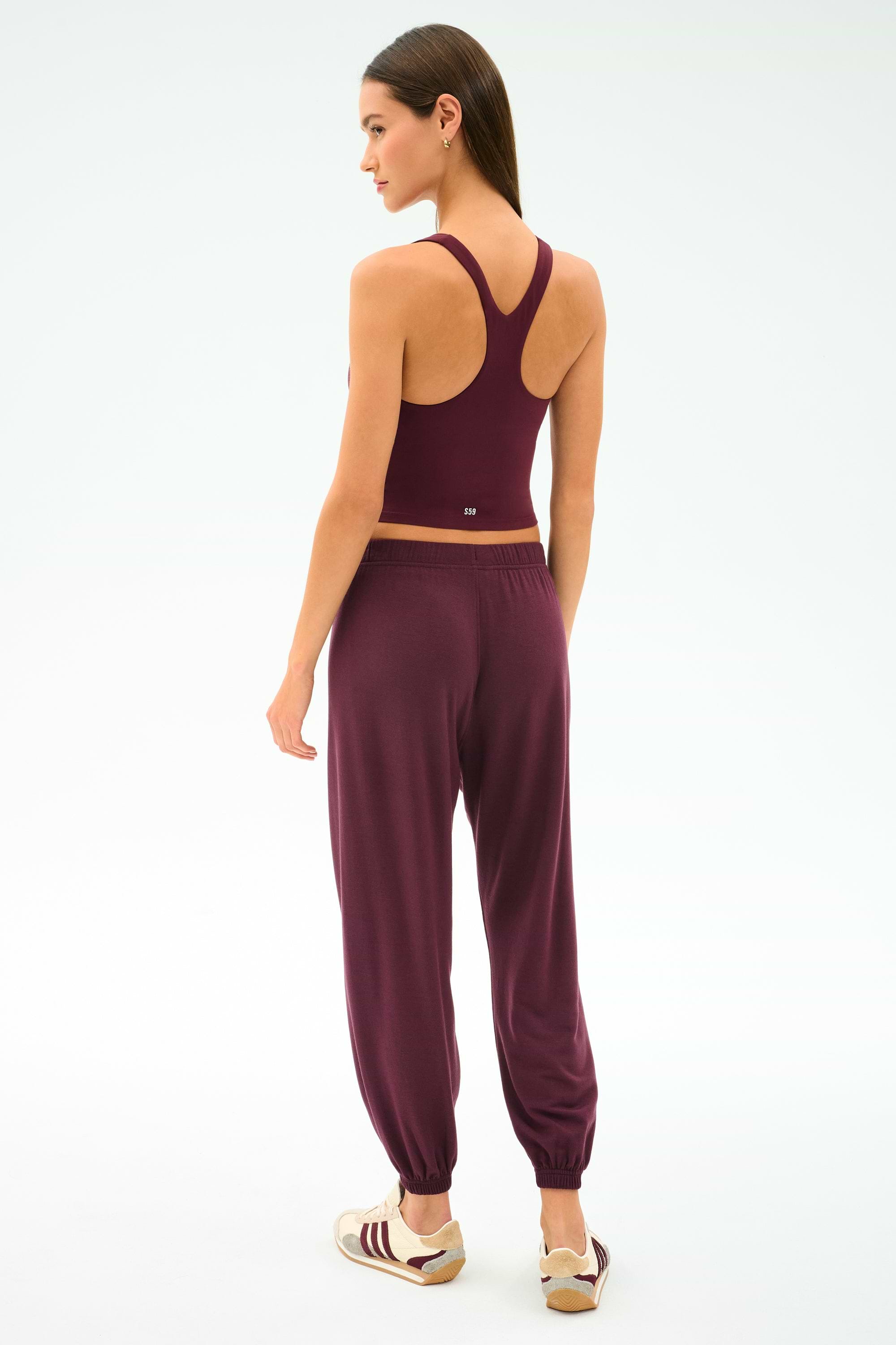A woman faces away, wearing the SPLITS59 Ella Airweight Tank in Dark Cherry/Valencia, made from stretchy nylon spandex, paired with jogger pants and sneakers against a plain white background.