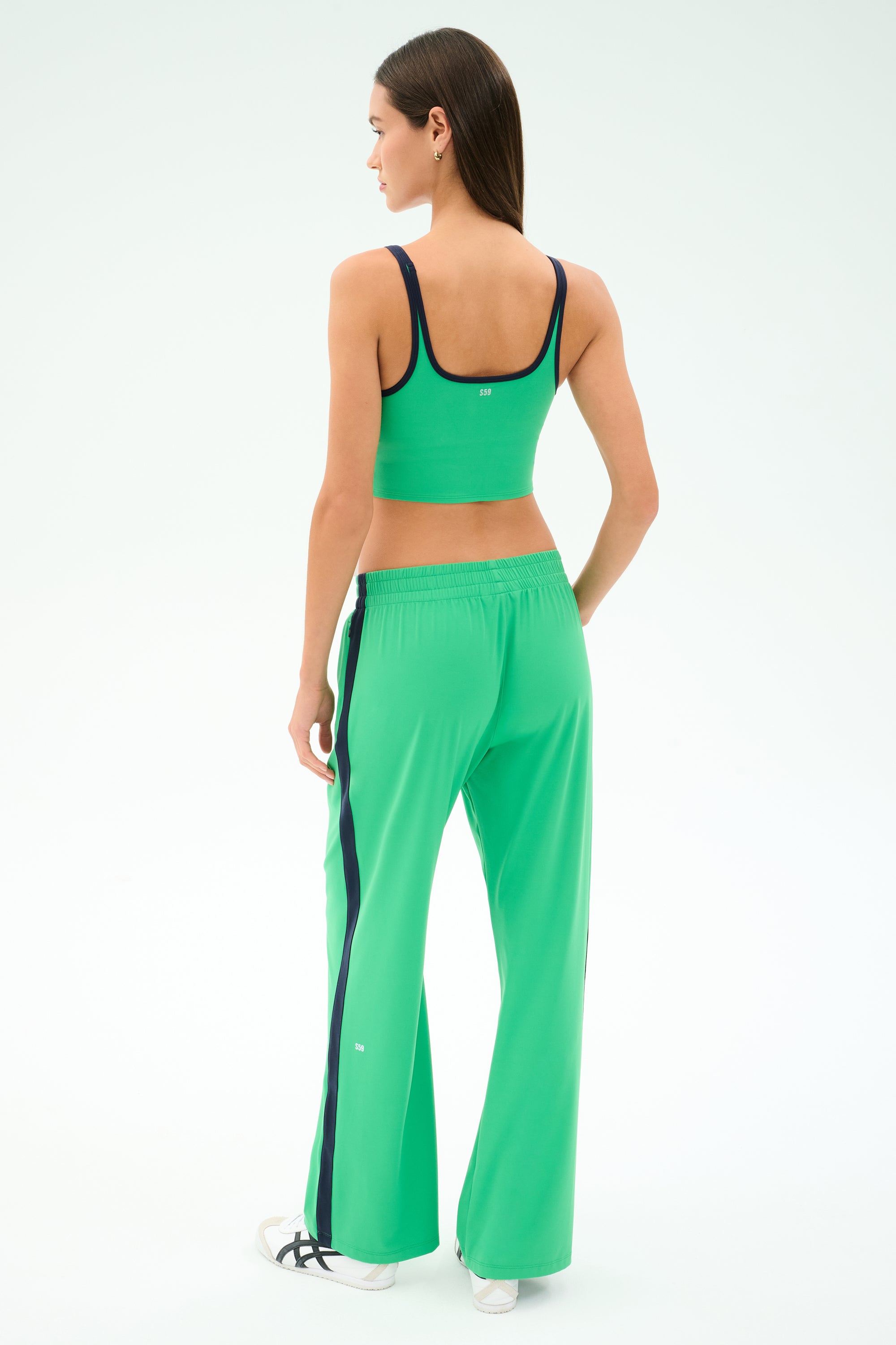 A woman faces away, wearing the SPLITS59 Cait Rigor Bralette in Grass/Indigo with matching green track pants featuring black stripes and white sneakers, set against a light background.