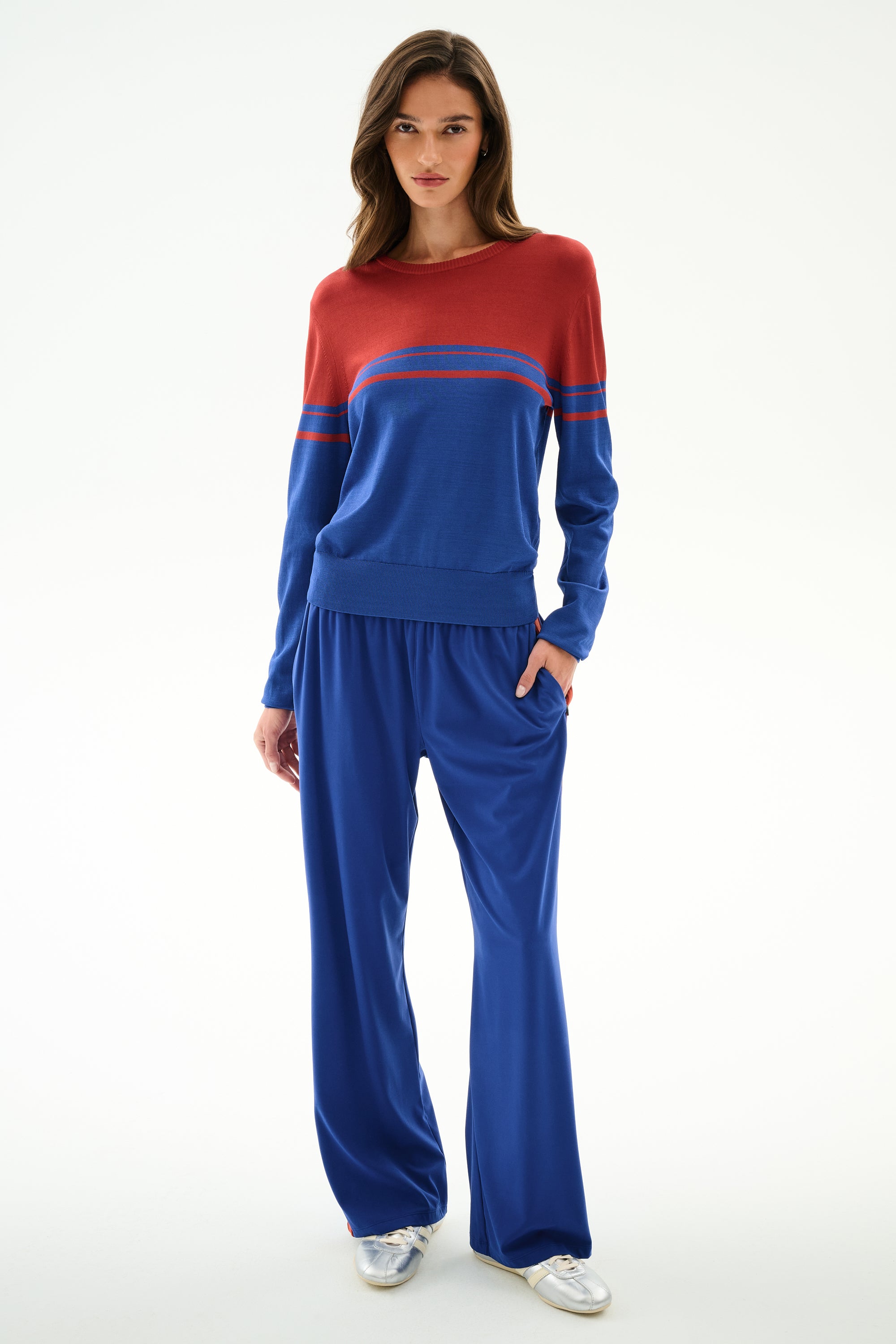 Woman stands against a white background in the SPLITS59 Anna Knit Pullover - Cobalt/Terracotta, blue wide-leg pants, and silver sneakers, left hand in her pocket.