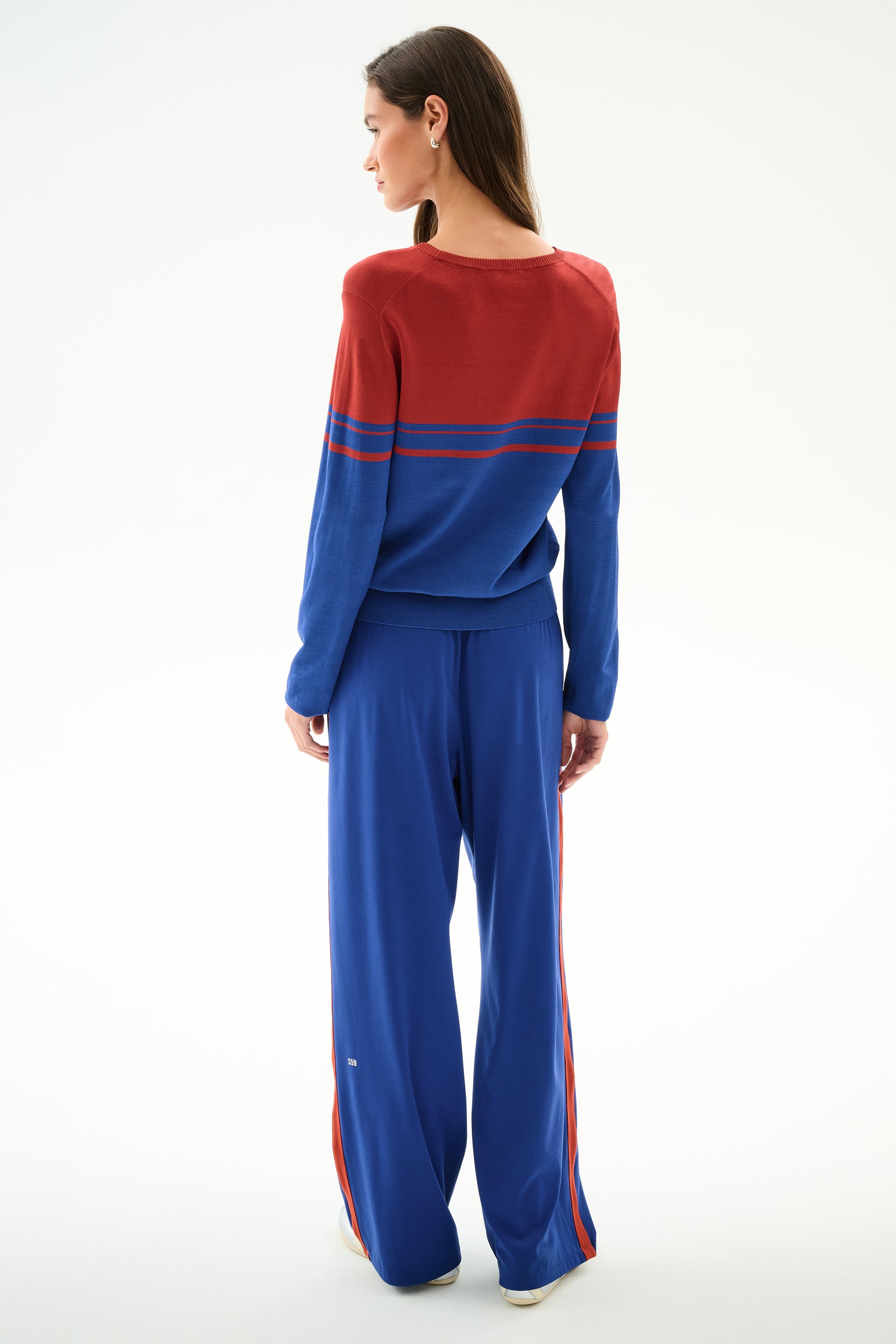 A woman faces away wearing the SPLITS59 Anna Knit Pullover - Cobalt/Terracotta, paired with coordinating wide-leg blue pants with red stripes, against a plain background.
