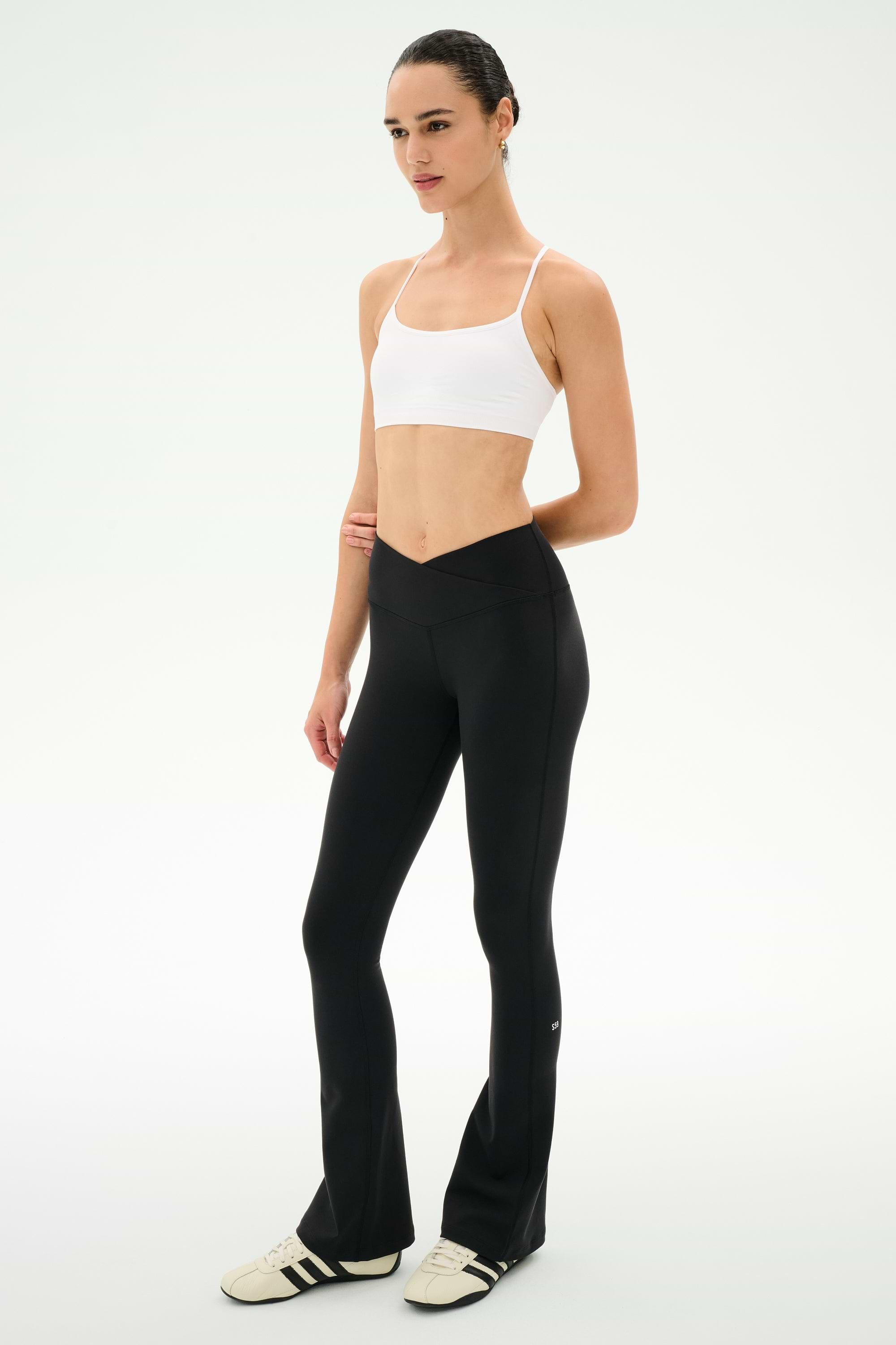 A person stands on a white background, facing slightly to the right, wearing a white sports bra, SPLITS59 Raquel Crossover Supplex Flare leggings in black made from 4-way stretch Supplex, and white sneakers.
