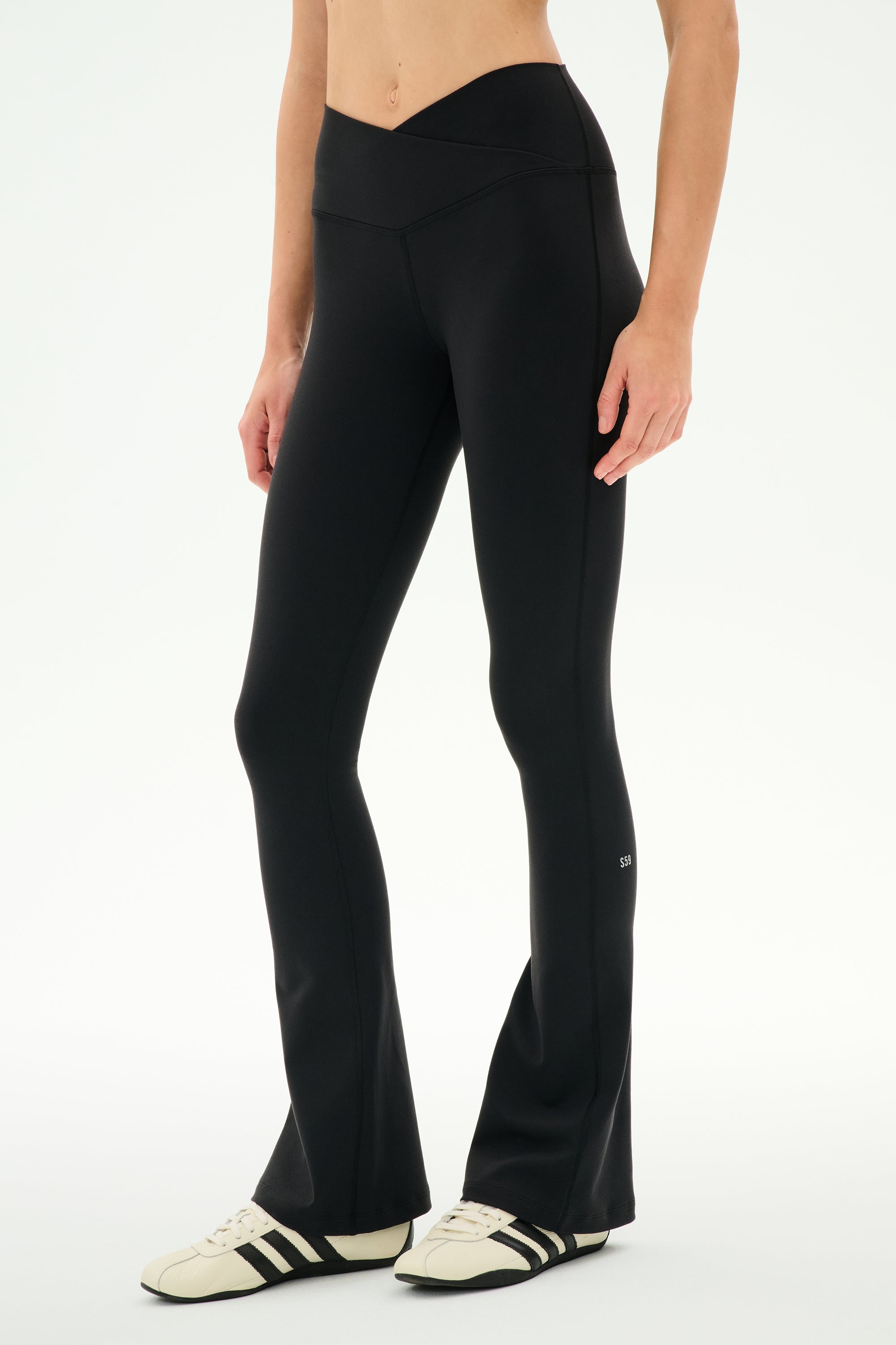 Wearing the SPLITS59 Raquel Crossover Supplex Flare in Black—high-waisted, flared leggings crafted from 4-way stretch Supplex—a person stands against a plain white background in white sneakers with black stripes. Perfect for yoga, these crossover waist leggings deliver both comfort and style.