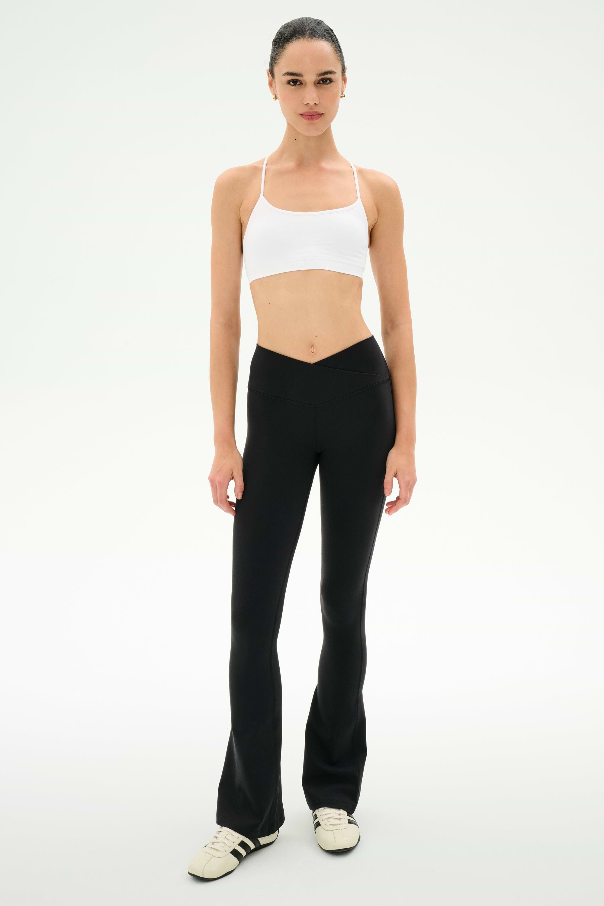 A woman stands facing forward on a plain white background, wearing a white sports bra, white sneakers, and the SPLITS59 Raquel Crossover Supplex Flare leggings in black, featuring a V-shaped crossover waistband and made from 4-way stretch Supplex fabric.