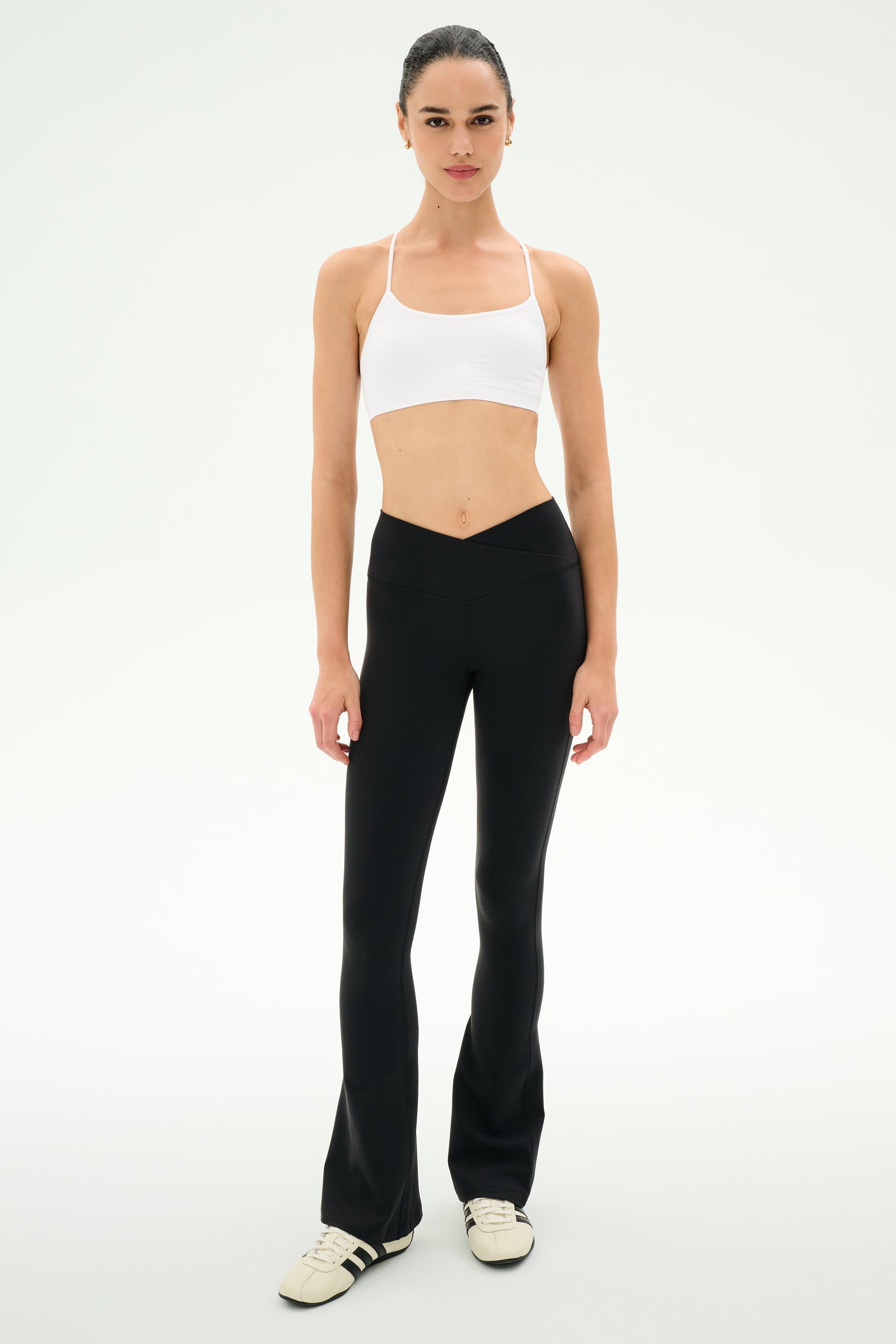 A woman stands facing forward on a plain white background, wearing a white sports bra, white sneakers, and the SPLITS59 Raquel Crossover Supplex Flare leggings in black, featuring a V-shaped crossover waistband and made from 4-way stretch Supplex fabric.