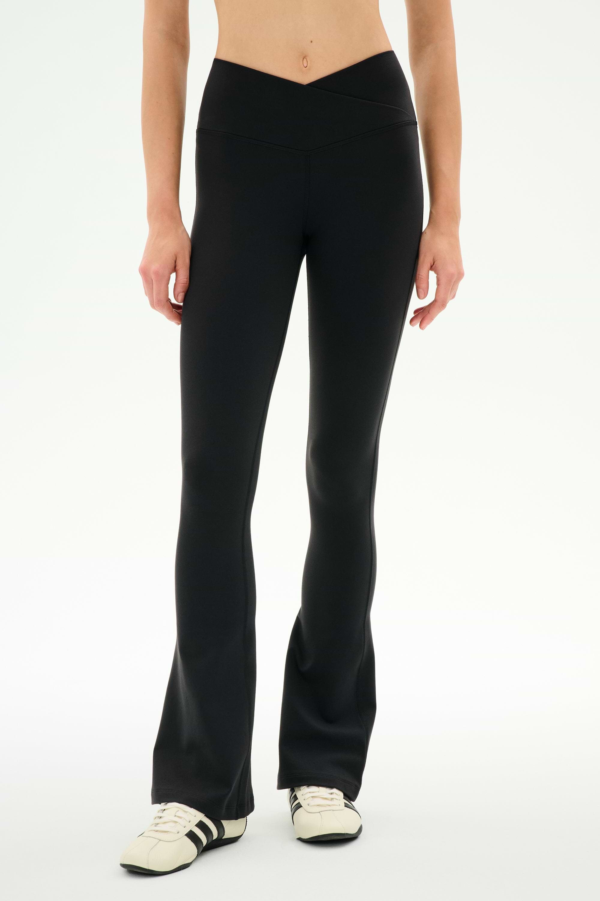 A person wearing the SPLITS59 Raquel Crossover Supplex Flare leggings in black and white sneakers stands against a plain white background, with only their lower body visible.