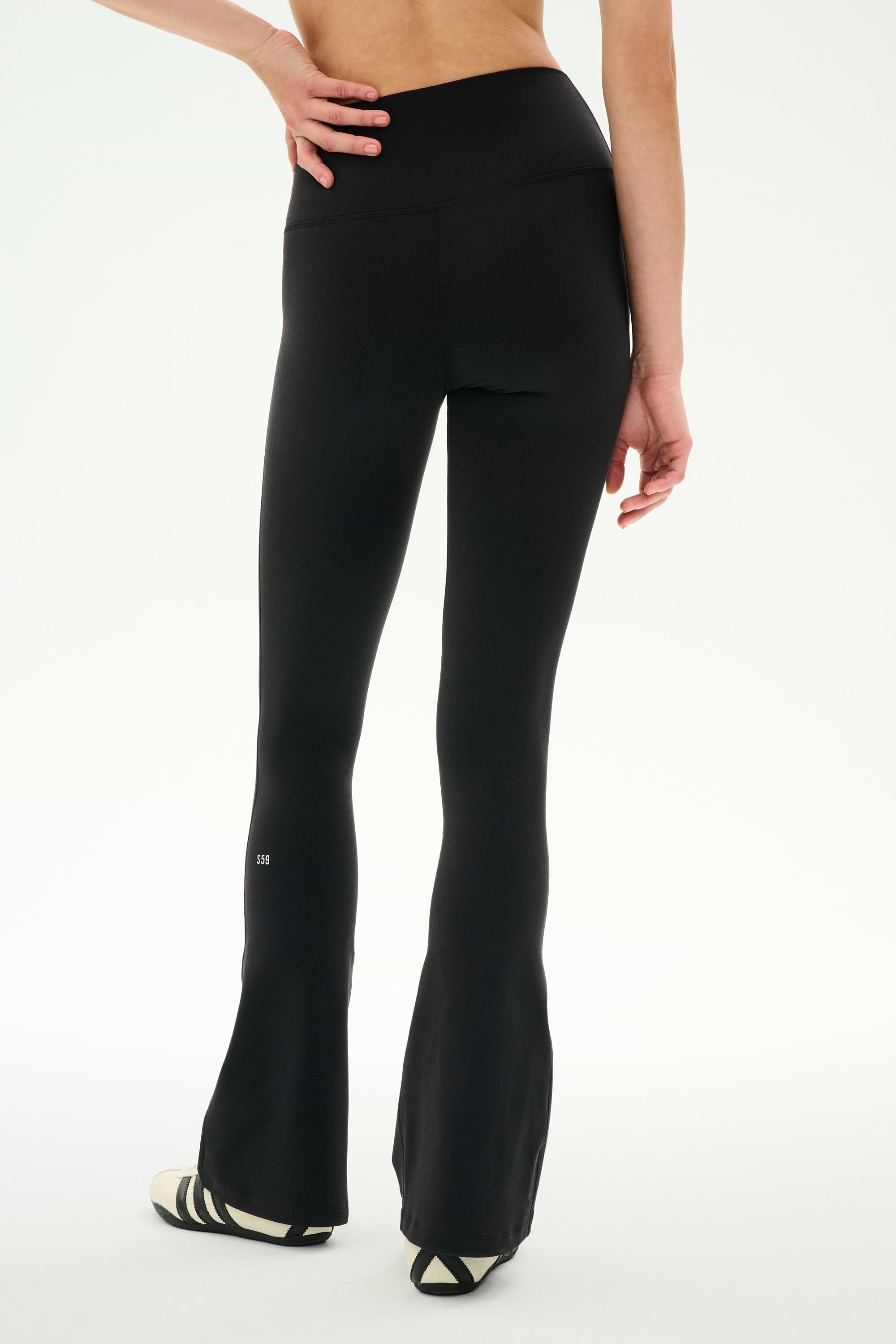 A person stands with one hand on their hip, shown from the back, wearing SPLITS59’s Raquel Crossover Supplex Flare leggings in black—featuring a flattering crossover waist and high-rise fit—paired with black-and-white sneakers against a plain white background; perfect for yoga sessions or comfortable everyday wear.