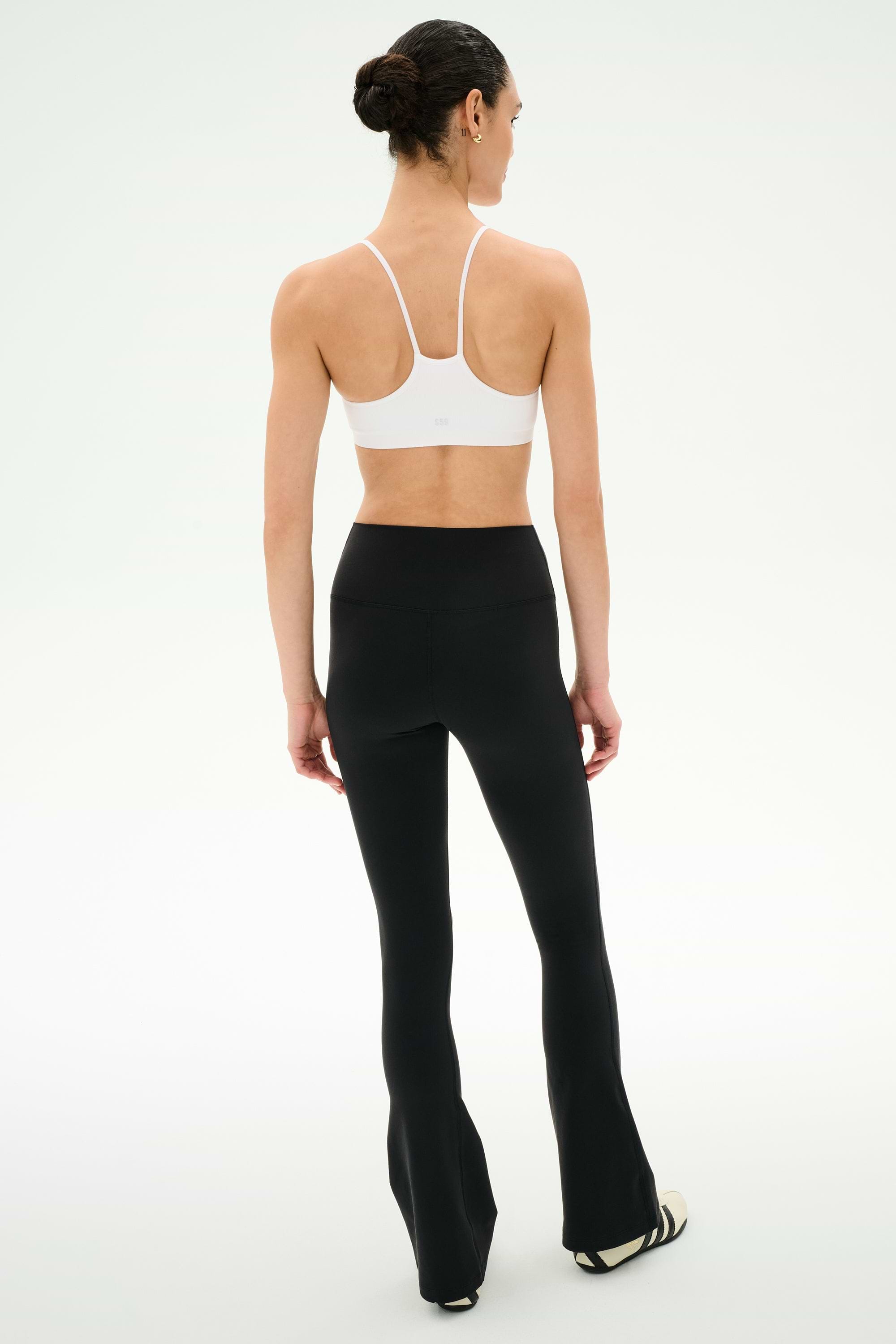 A person stands with their back to the camera, wearing the SPLITS59 Raquel Crossover Supplex Flare in Black, featuring a high-waisted crossover waistband made from 4-way stretch Supplex fabric, paired with a white sports bra and black and white shoes against a plain white background.