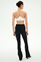 A person stands with their back to the camera, wearing the SPLITS59 Raquel Crossover Supplex Flare in Black, featuring a high-waisted crossover waistband made from 4-way stretch Supplex fabric, paired with a white sports bra and black and white shoes against a plain white background.