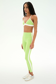 A woman stands in profile against a plain white background, wearing the SPLITS59 Sage Rigor Bra in Limeade/White with matching leggings and white sneakers.