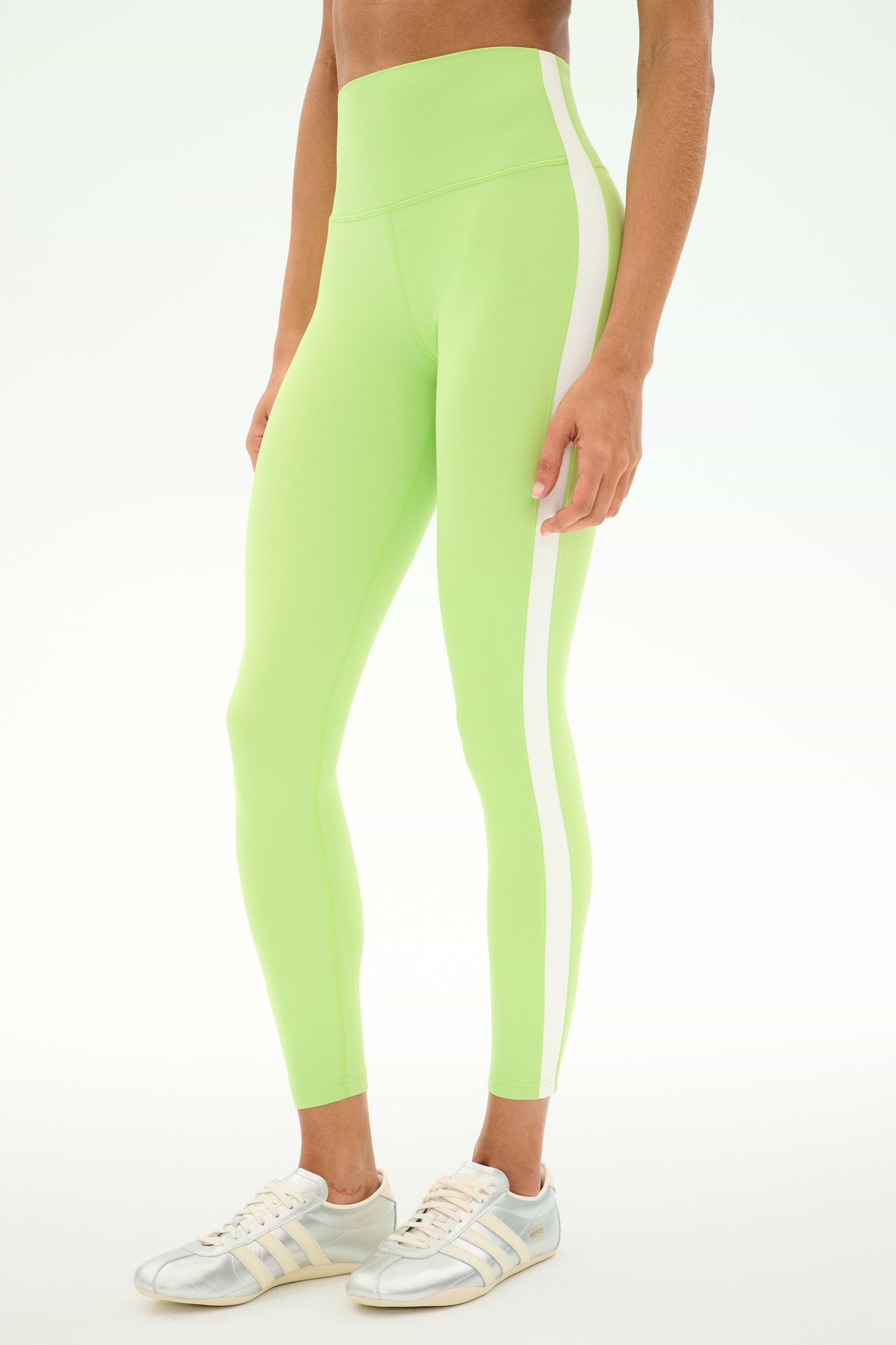 A person stands against a plain background wearing the SPLITS59 Clare High Waist Rigor 7/8 leggings in Limeade/White—a high-waisted style crafted from a soft nylon-spandex blend with a white side stripe—paired with white sneakers.