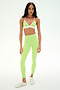 A woman stands facing forward against a plain light background wearing the SPLITS59 Clare High Waist Rigor 7/8 leggings and matching sports bra set in Limeade/White, crafted from a soft nylon-spandex blend, and paired with white sneakers.