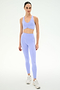 A woman faces forward in the SPLITS59 Airweight Bra with Ruching in Purple Haze and matching high-waisted leggings, paired with white sneakers, against a plain white background.