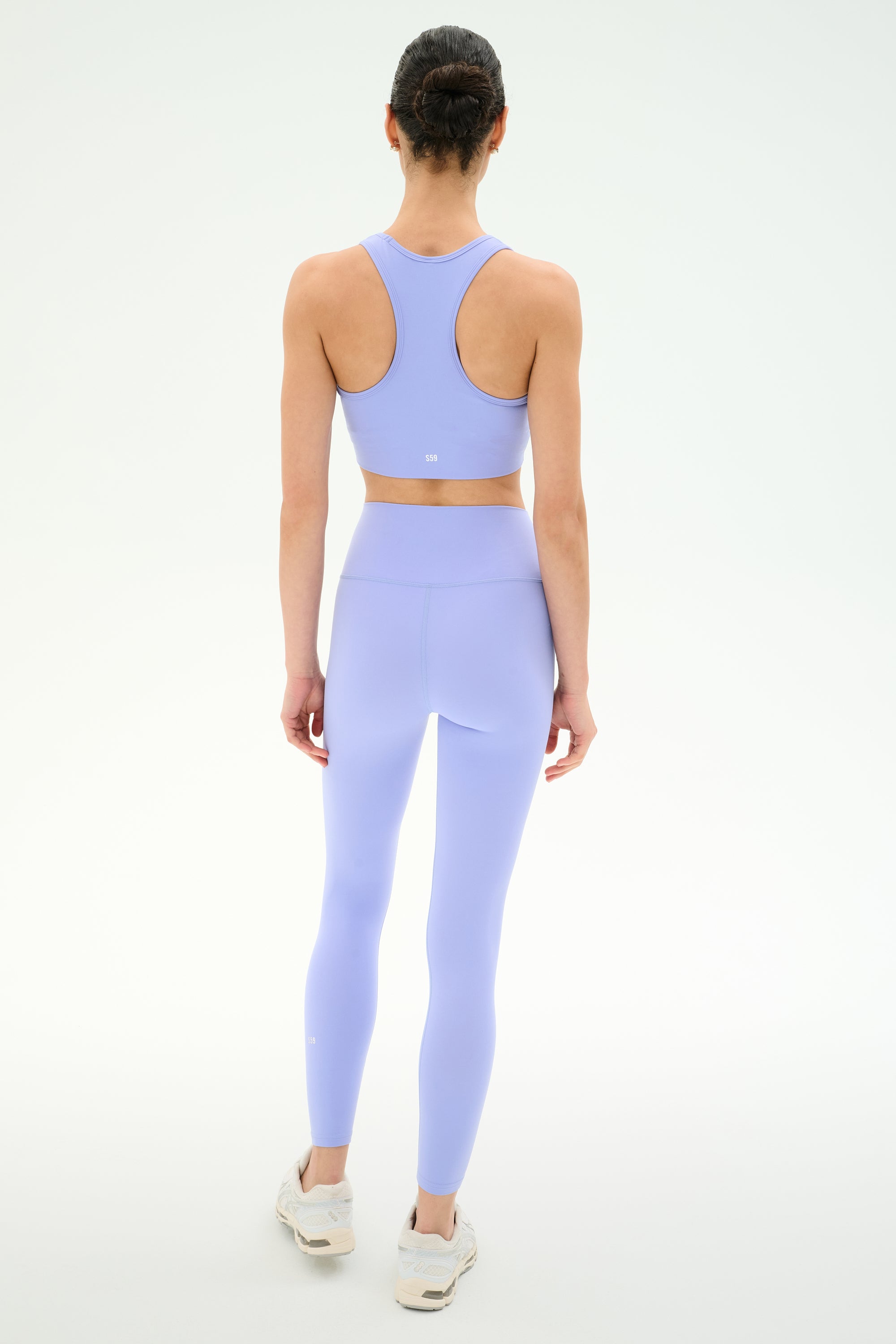 A person faces away, wearing SPLITS59's Airweight High Waist 26" leggings in Purple Haze with a matching sports bra and white sneakers, set against a plain white background.