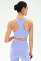 A woman with dark hair in a bun wears the SPLITS59 Airweight Bra with Ruching in Purple Haze and matching high-waisted leggings, facing away from the camera against a plain background.