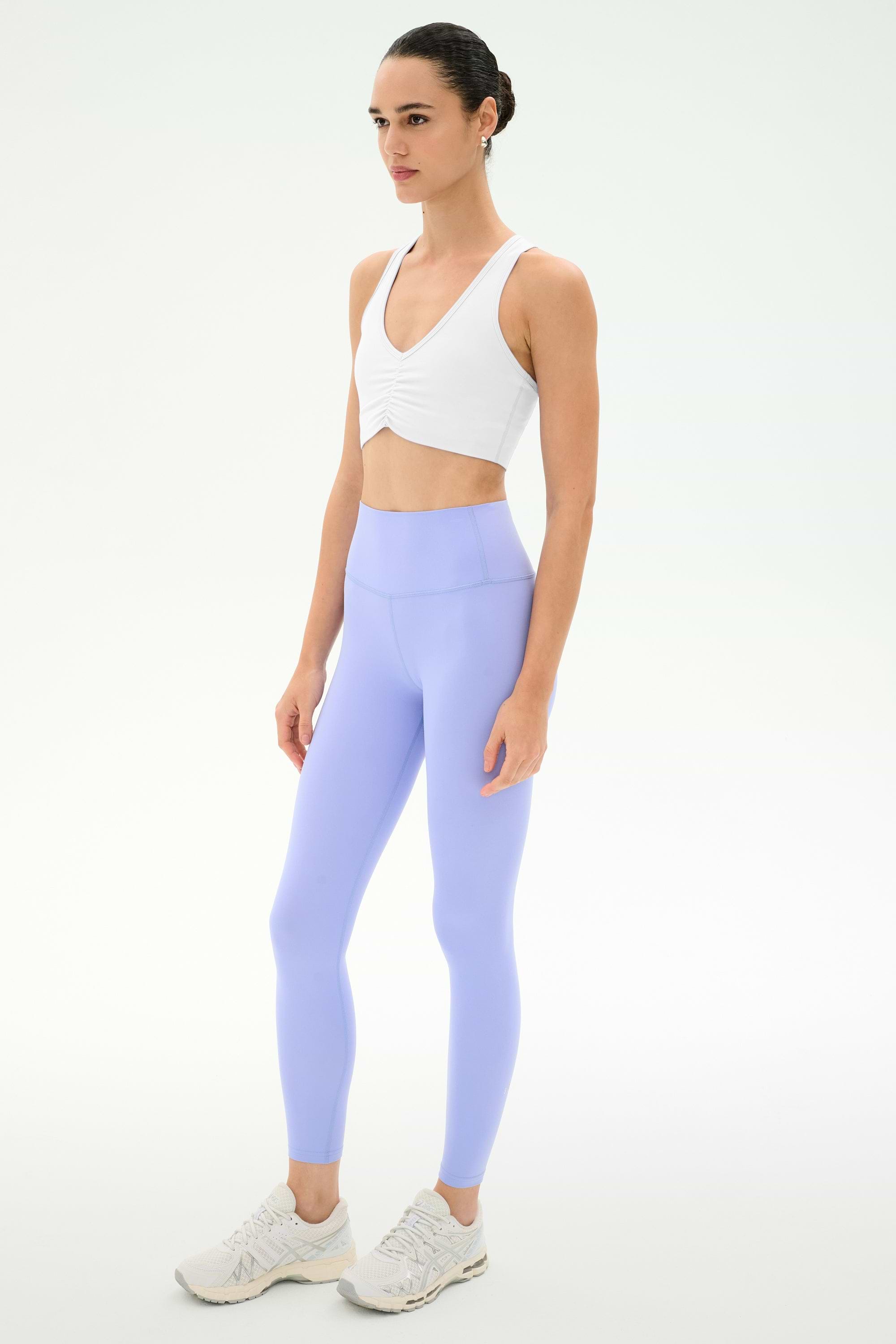 A person wears the SPLITS59 Airweight Bra with Ruching in white, light purple leggings, and white sneakers, standing against a plain light background.