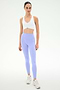 A woman faces forward in a SPLITS59 Airweight Bra with Ruching in white, paired with light purple high-waisted leggings and white sneakers, against a plain light background.