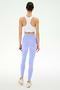 A woman faces away, wearing the SPLITS59 Airweight Bra with Ruching in white, paired with light purple leggings and white sneakers, set against a plain white background.