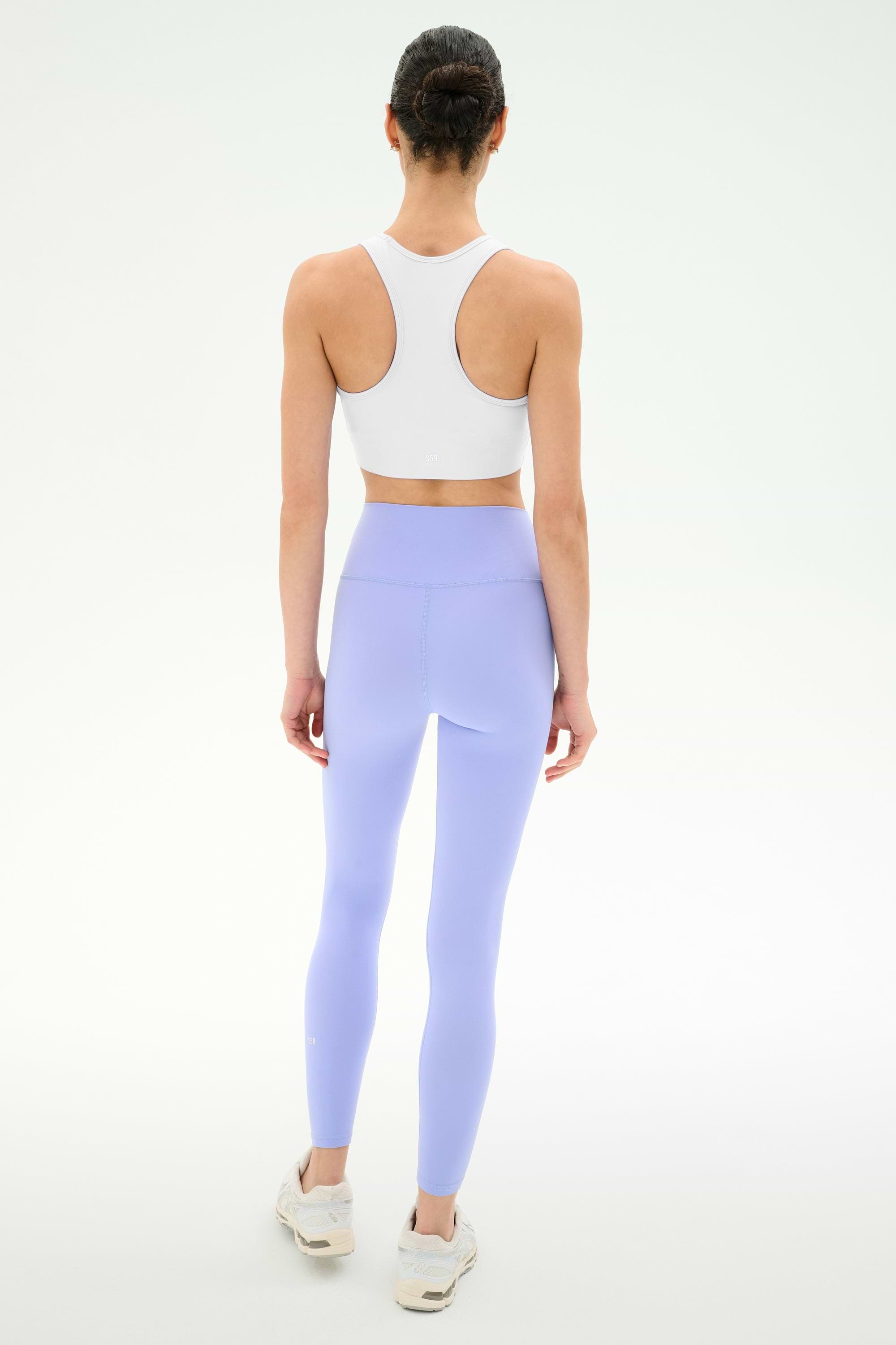 A woman faces away, wearing the SPLITS59 Airweight Bra with Ruching in white, paired with light purple leggings and white sneakers, set against a plain white background.