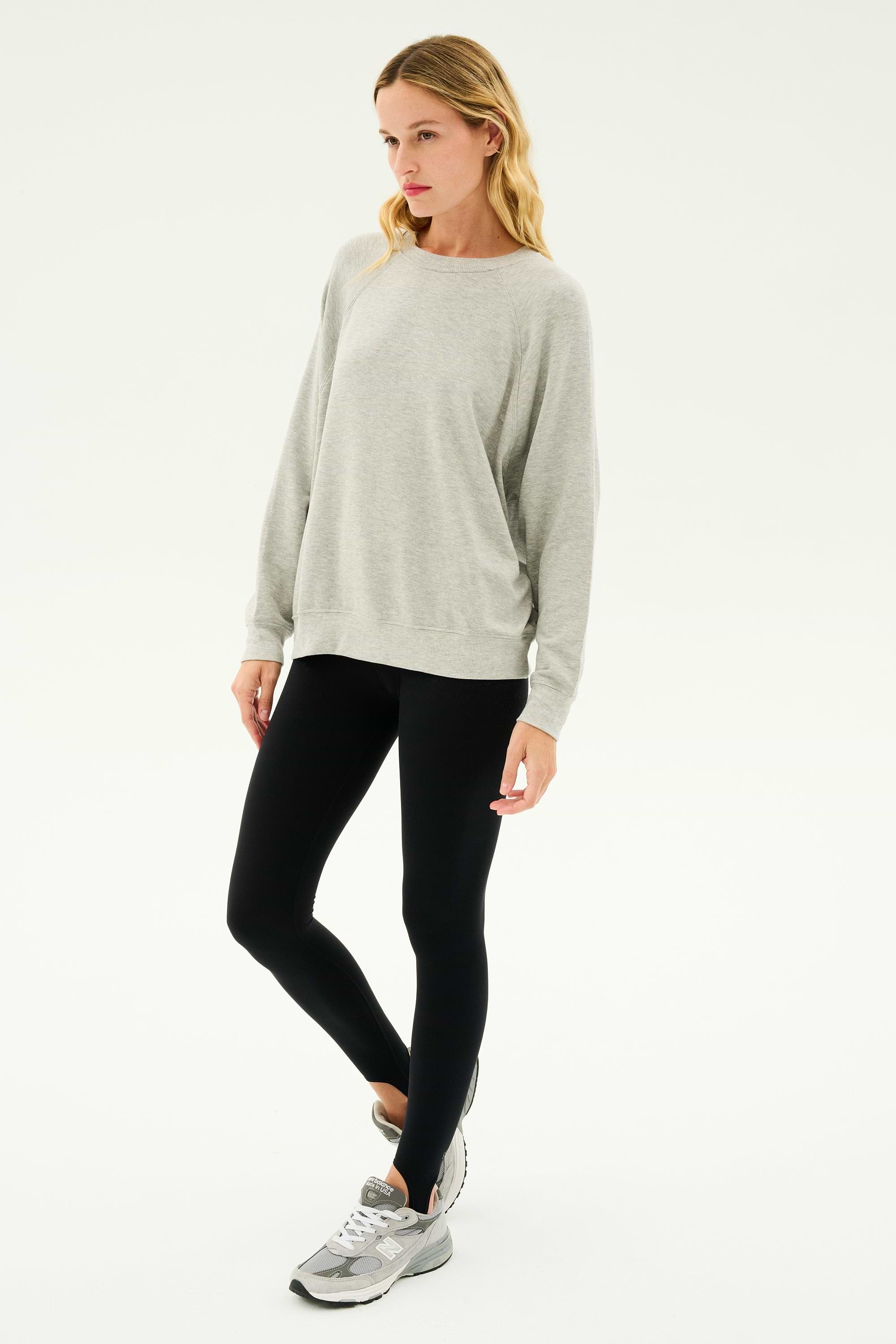 A person wearing an Andie Fleece Sweatshirt in Heather Grey by SPLITS59, black leggings, and grey sneakers stands against a plain white background.