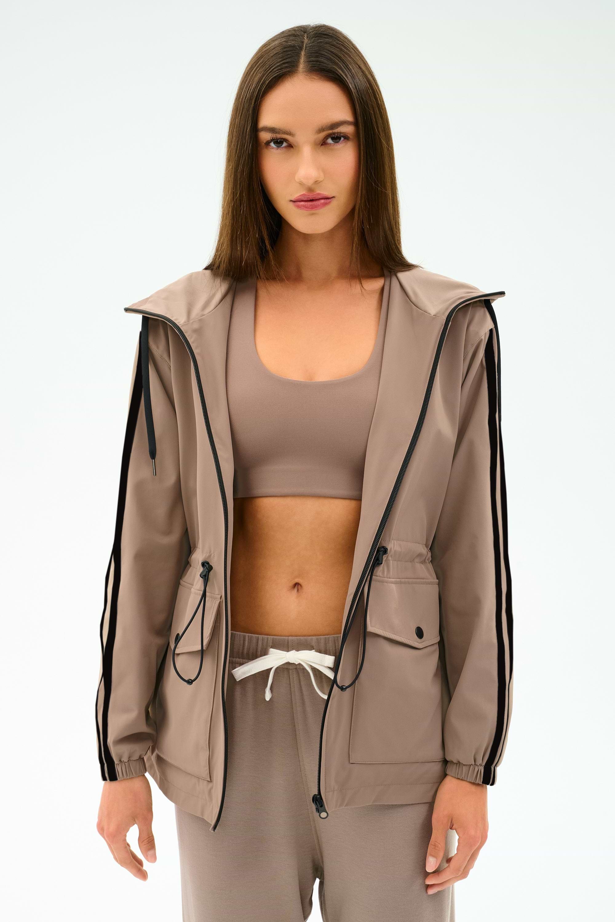 A woman with straight brown hair wears the SPLITS59 Reggie Water Repellent Jacket in Latte/Black, styled with matching taupe sportswear, against a plain white background.