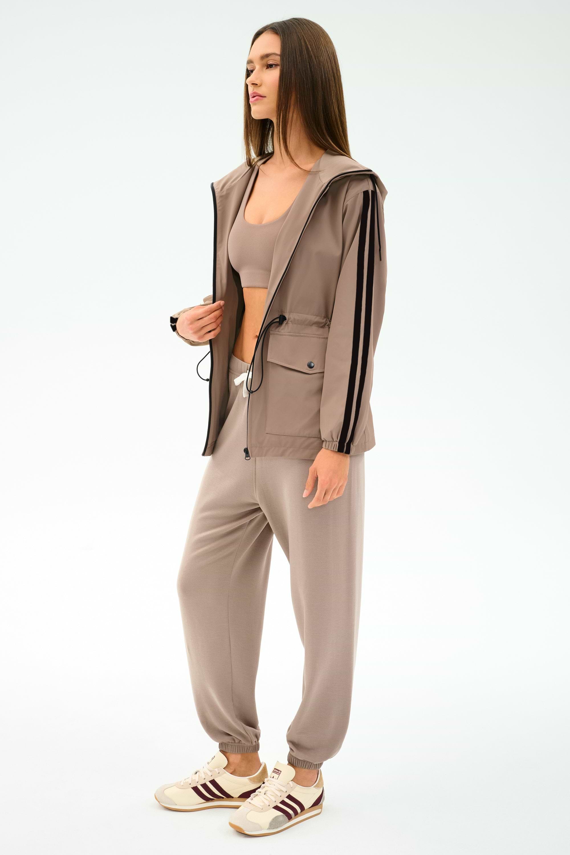Woman wearing the SPLITS59 Reggie Water Repellent Jacket in Latte/Black, styled with matching joggers, a crop top, and beige sneakers against a plain light background.