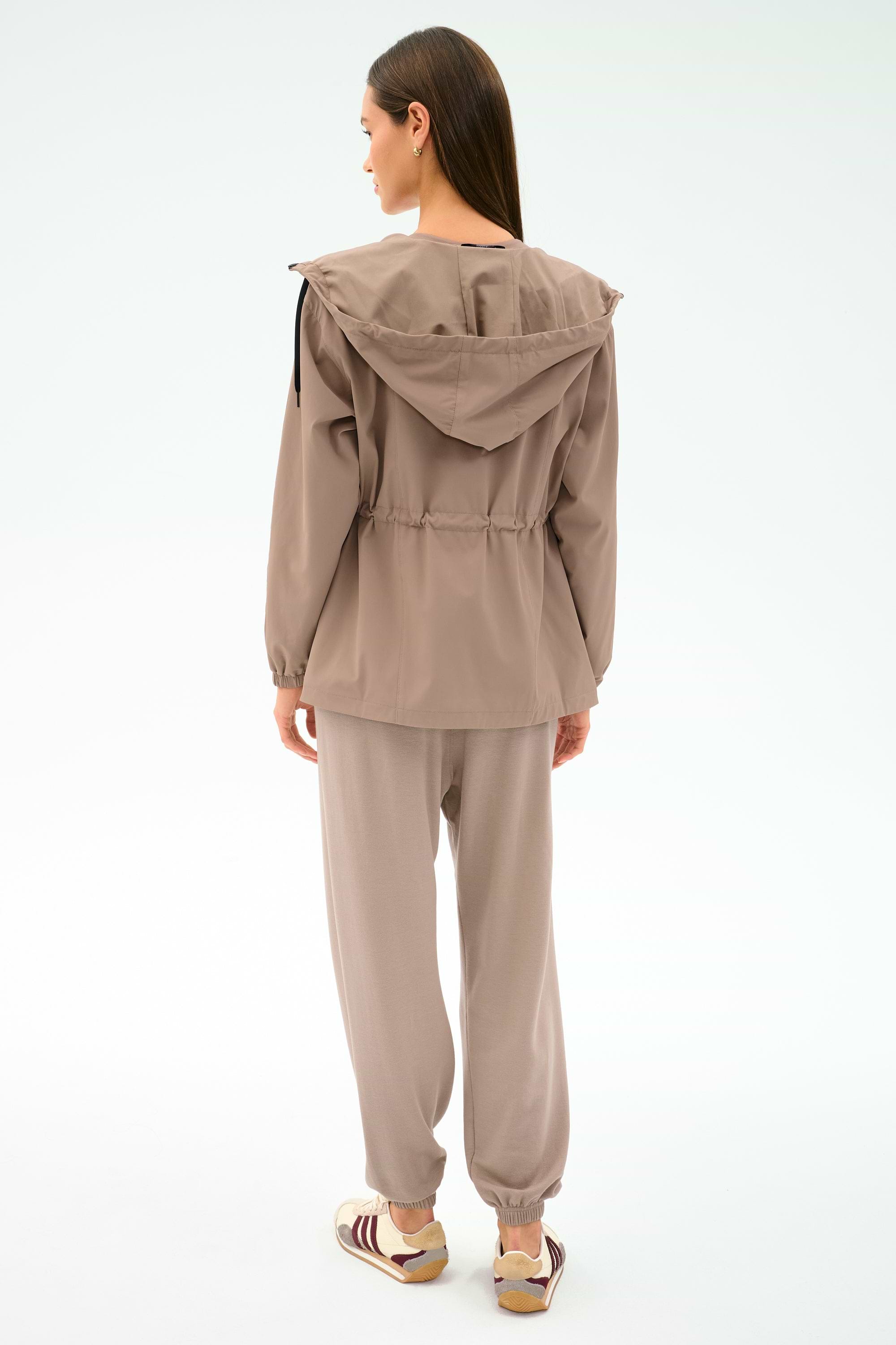 A woman stands with her back to the camera, wearing the SPLITS59 Reggie Water Repellent Jacket in Latte/Black, paired with matching casual pants and beige-maroon sneakers against a light background.