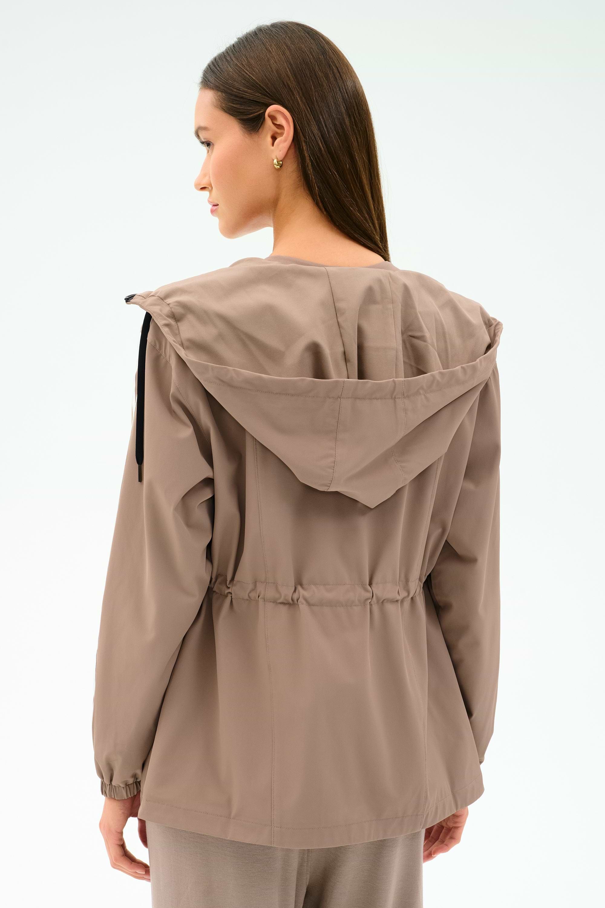A woman with long brown hair is seen from the back wearing the SPLITS59 Reggie Water Repellent Jacket in Latte/Black, featuring a stretch fabric, hood, and drawstring waist against a plain light background.