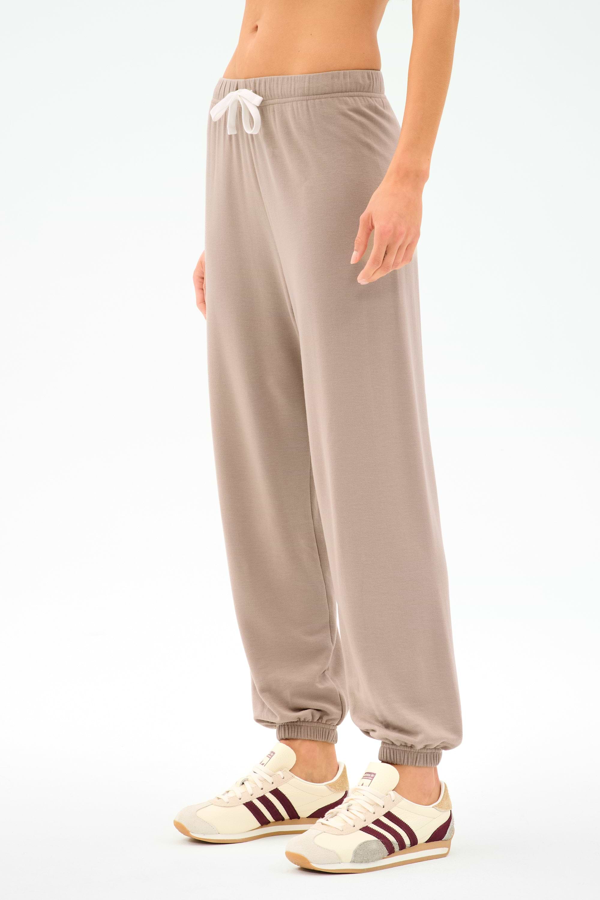 Person wearing the SPLITS59 Andie Oversized Fleece Sweatpant in Latte, featuring a white drawstring and paired with beige sneakers with maroon stripes, perfect for cool-weather workouts against a plain light background.