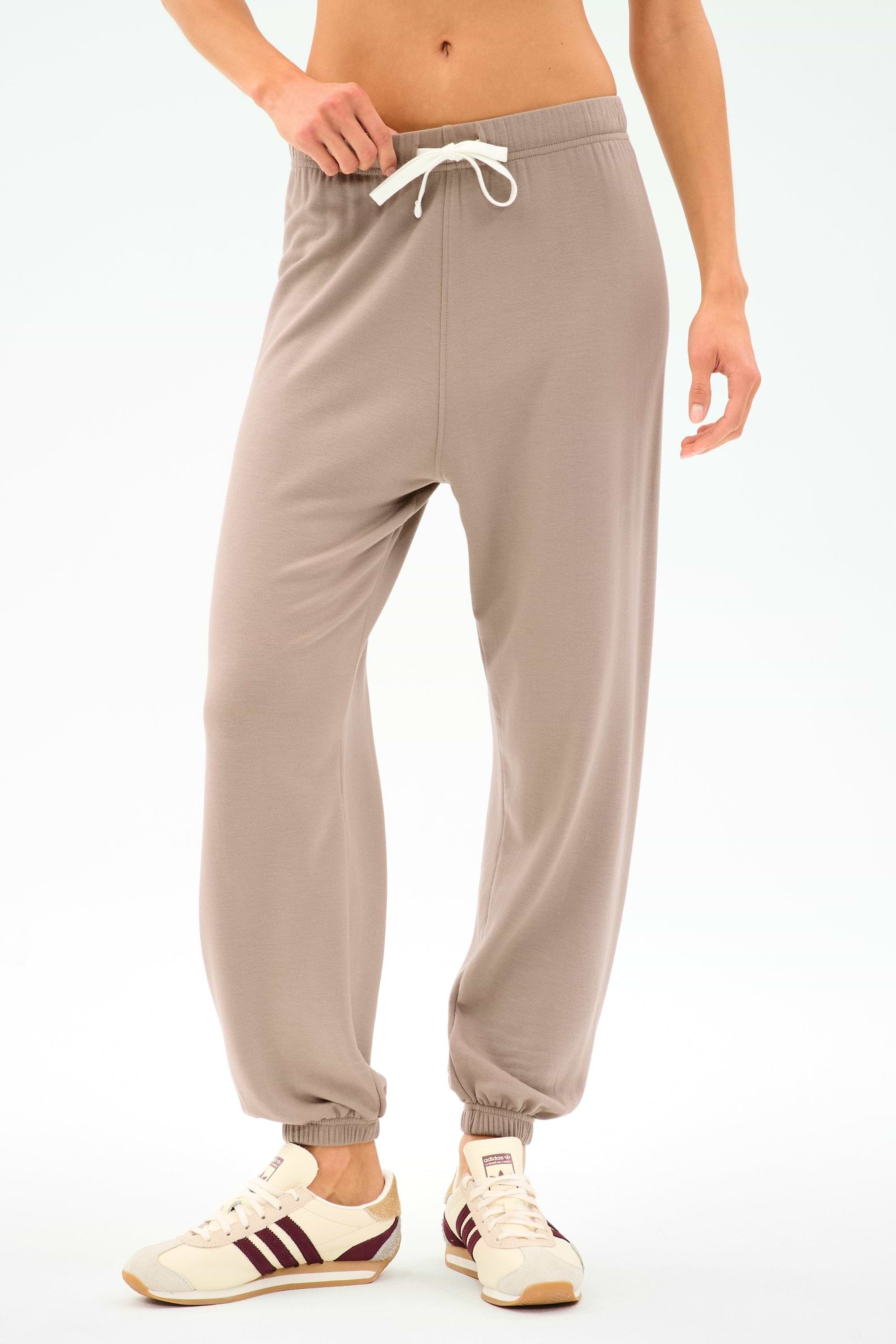 Wearing SPLITS59's Andie Oversized Fleece Sweatpant in Latte, featuring a drawstring waist and cuffed ankles, paired with white sneakers with maroon stripes—ideal for cool weather workouts or warm-ups against a plain background.