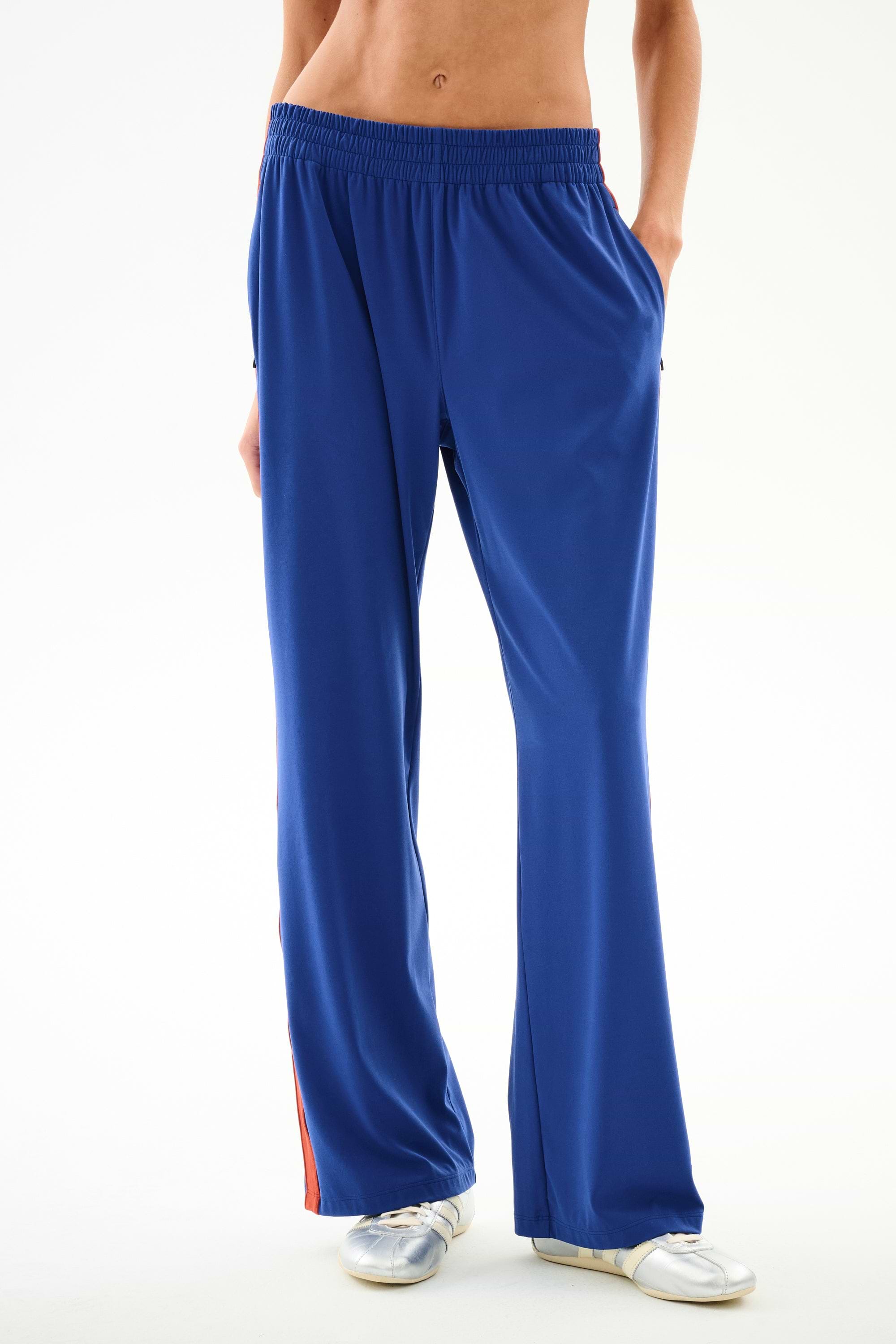 Model wears SPLITS59 Maxie Rigor Track Pant - Cobalt/Terracotta, with red side stripes, white sneakers, hands in pockets; shown waist down on white background.